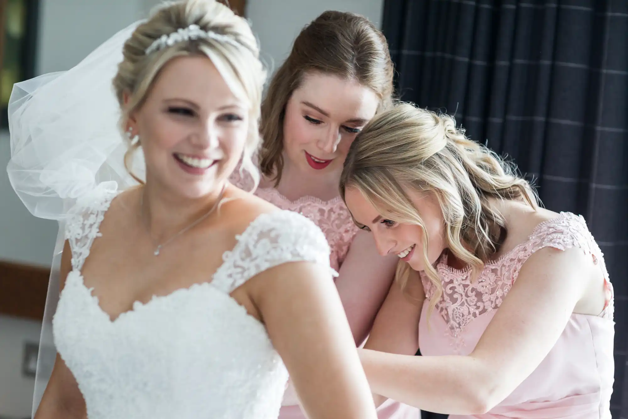 A bride in a white lace wedding dress is getting help adjusting her gown from two bridesmaids in pink dresses. All three women are smiling.