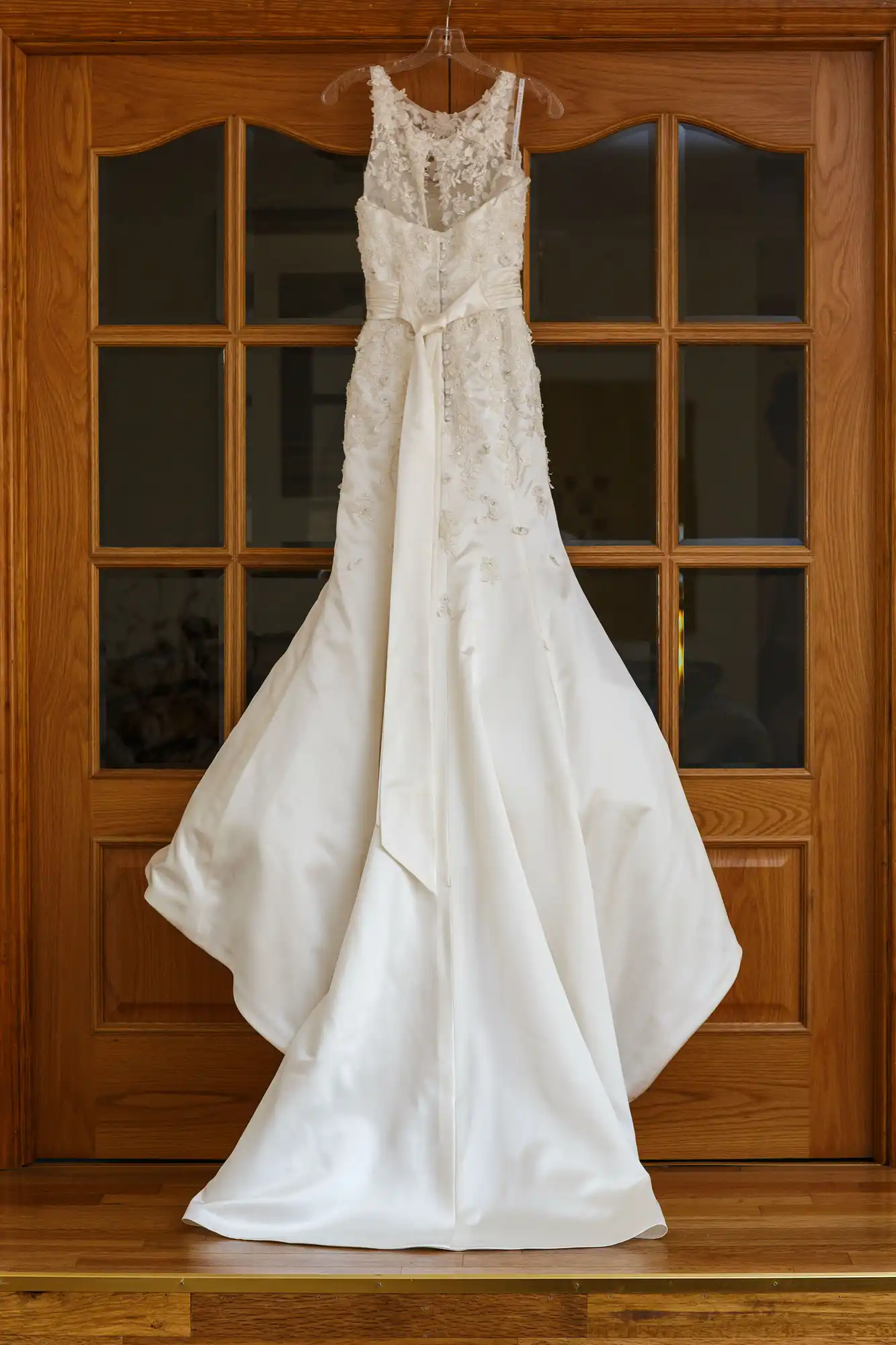 A white wedding dress with lace details hangs on a wooden door with glass panes.