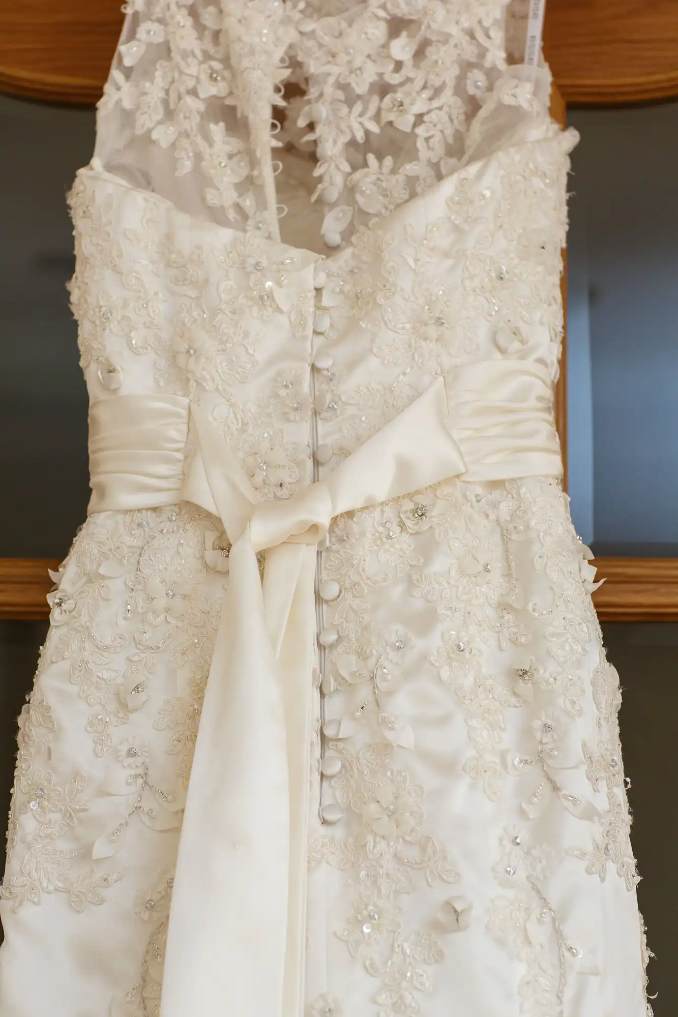 An intricately embroidered wedding dress with a satin bow hanging on a wooden door.