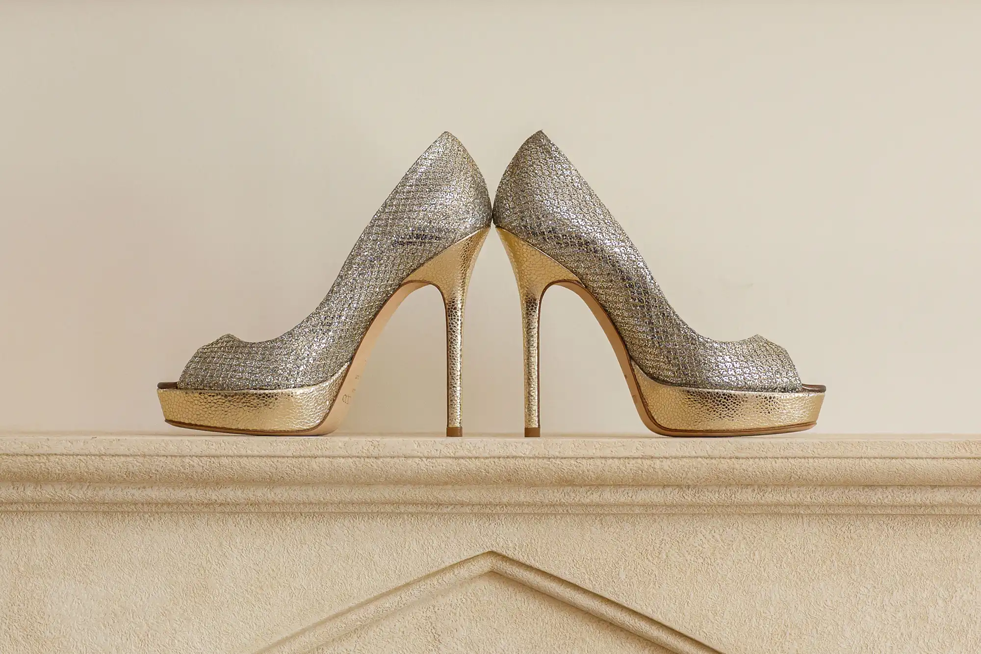 A pair of glittery silver high-heeled shoes with peep toes, displayed on a cream-colored ledge.