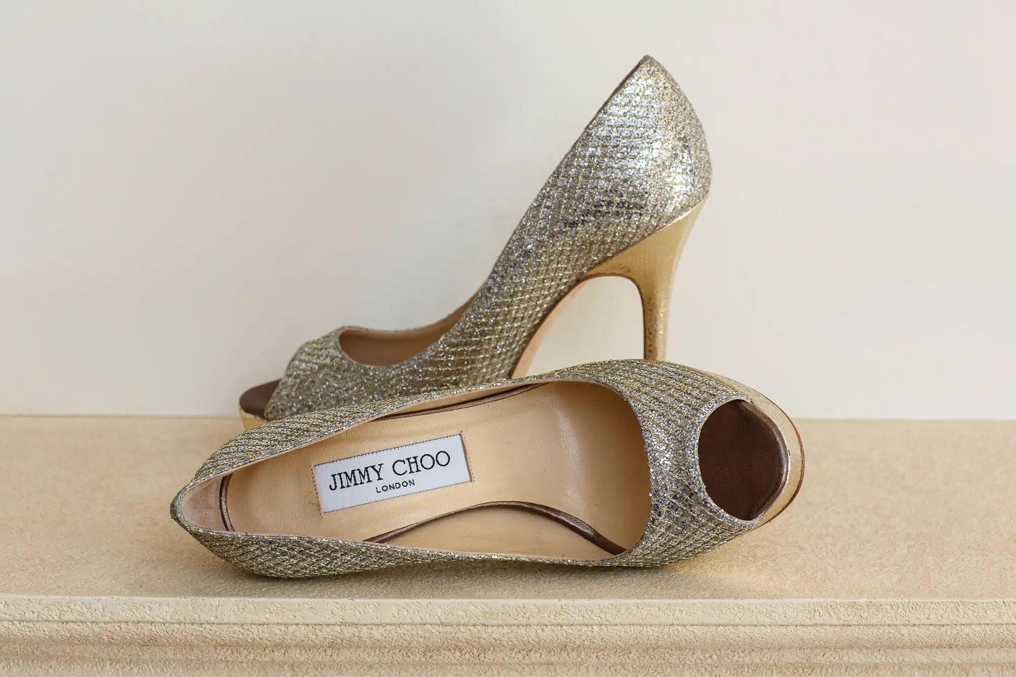 A pair of glittery gold jimmy choo high heels displayed on a beige surface against a neutral background.