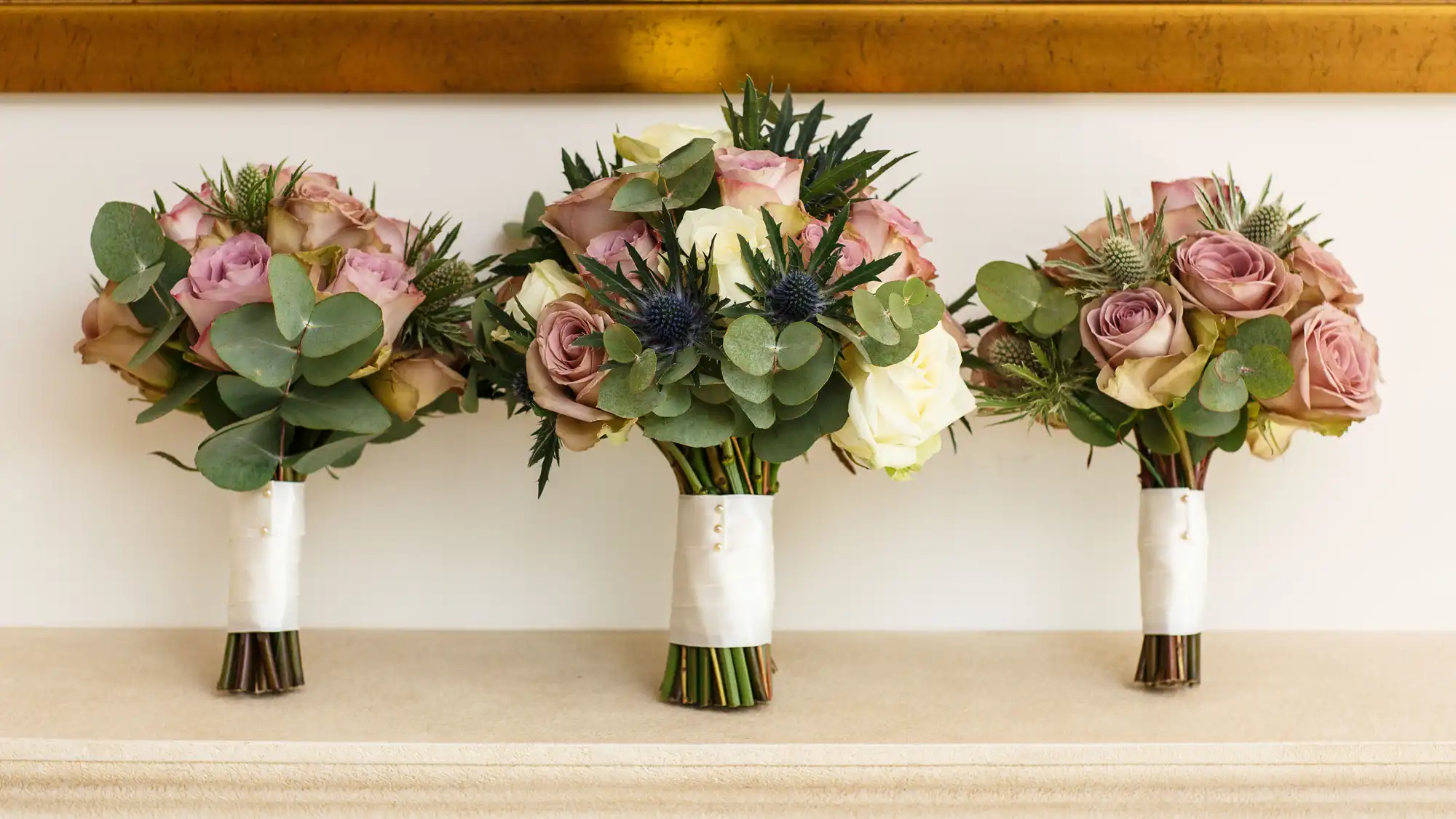 Three bridal bouquets with pink roses, white flowers, and blue thistles, wrapped in white ribbons, displayed on a table.