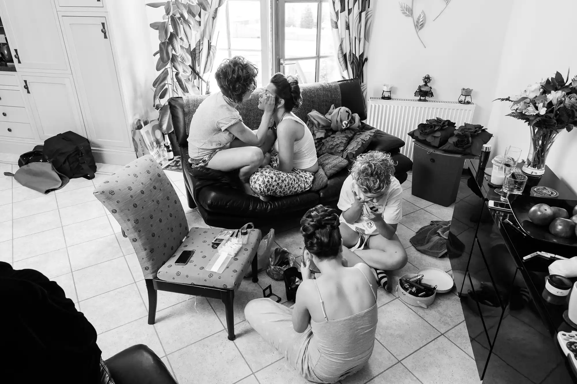 Black and white photo of four women in a living room, one getting her makeup done, others seated and chatting, with various items scattered around.
