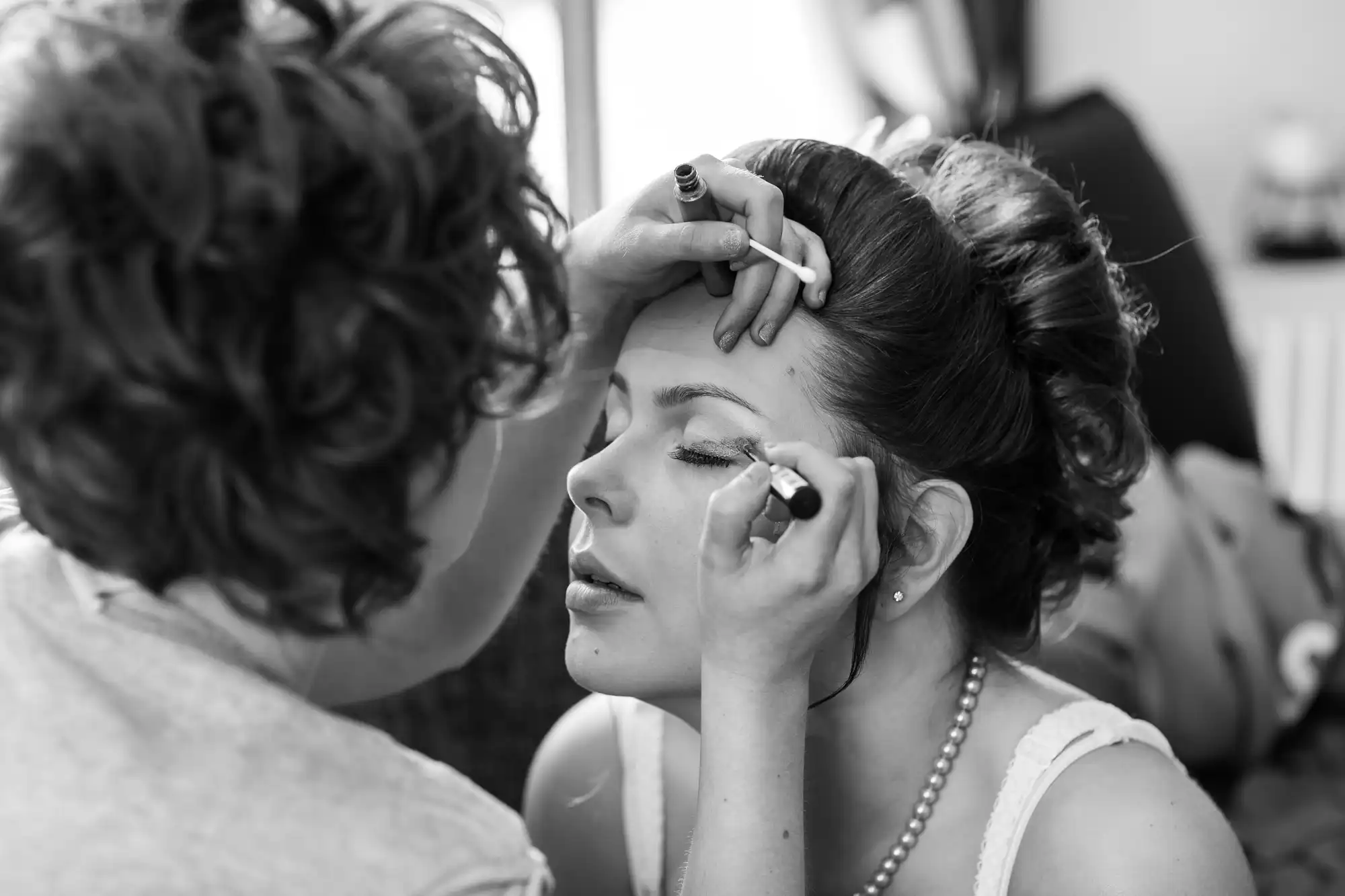 A woman applies eye makeup to another woman in a black and white photograph.