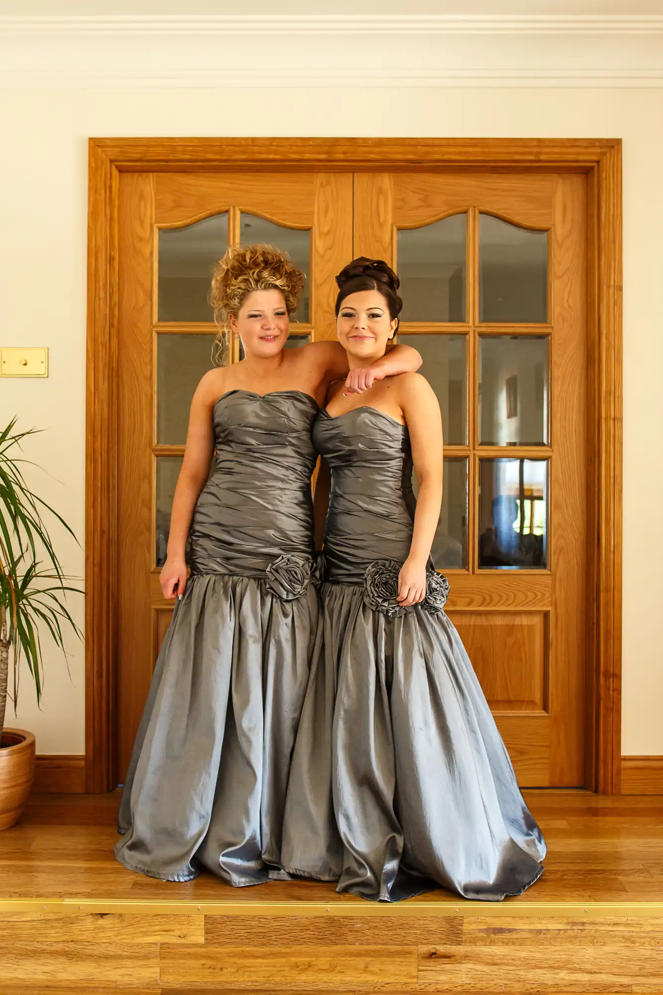 Two women in elegant gray ball gowns posing in front of wooden double doors.