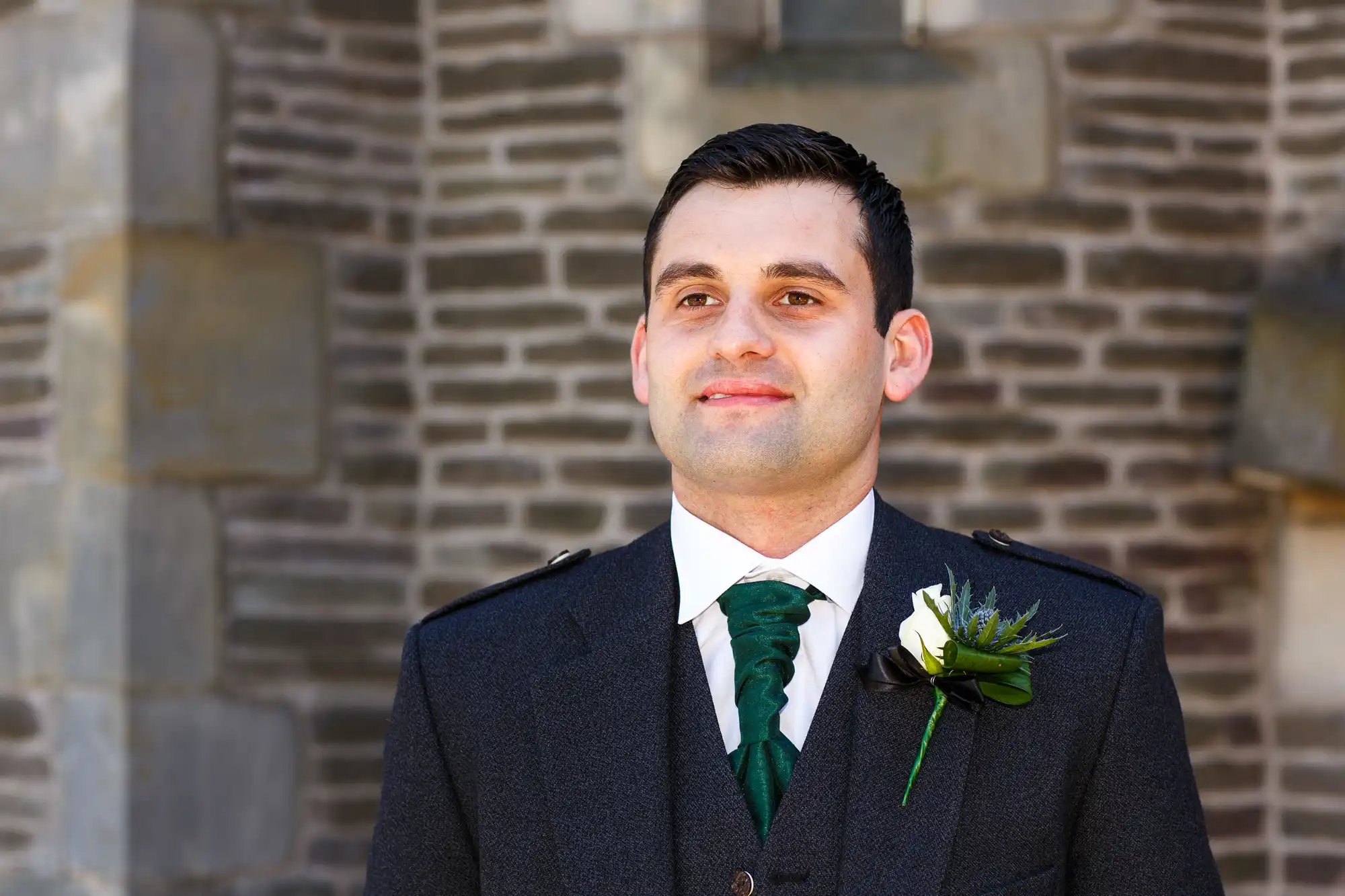 Man in a dark suit with green accents and a white rose boutonniere, smiling outside in front of a brick wall.