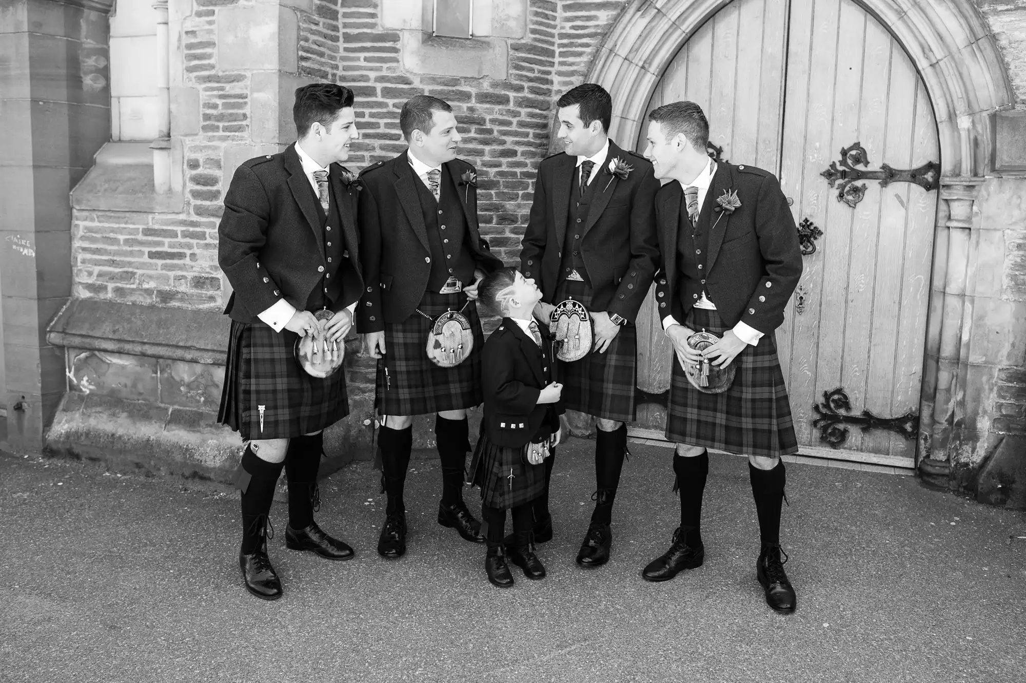 Four men in traditional scottish kilts and jackets gathered in conversation outside a historical stone building.