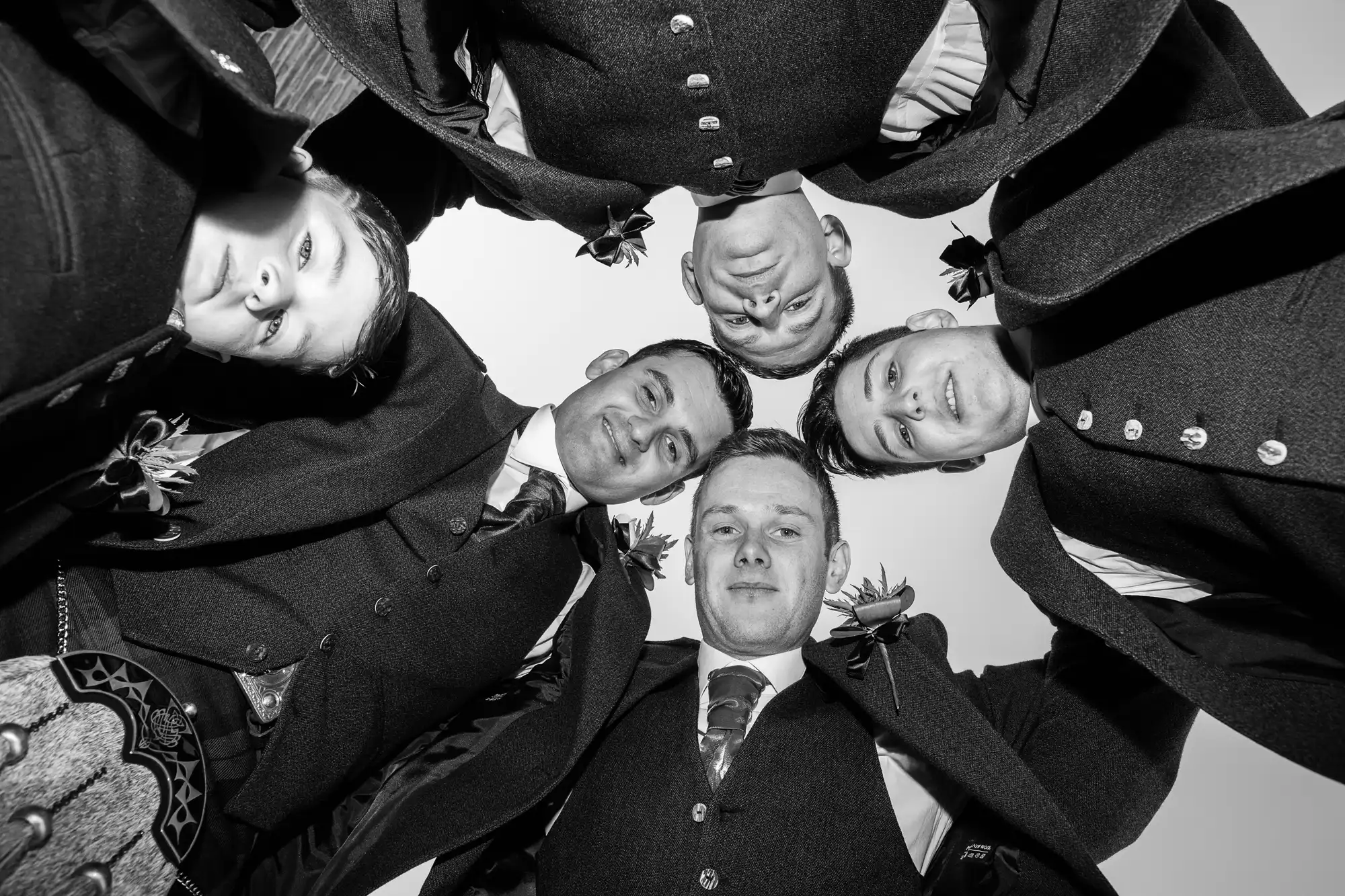 Five men in traditional suits with boutonnieres, viewed from below, forming a circle and looking down towards the camera.