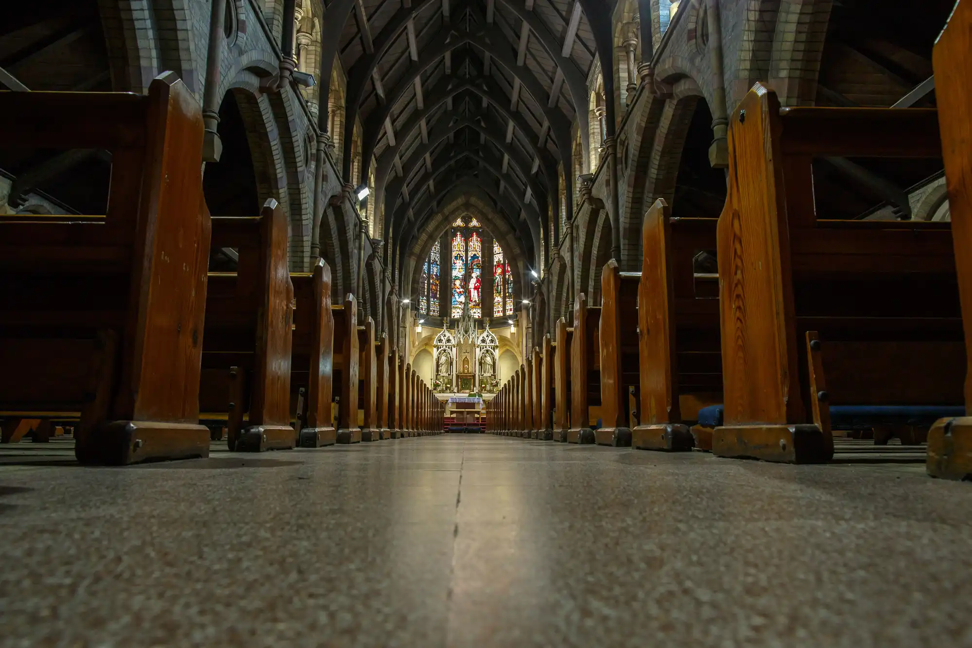 Low-angle view inside a cathedral, showcasing rows of wooden pews leading to an ornate altar and a stained glass window.
