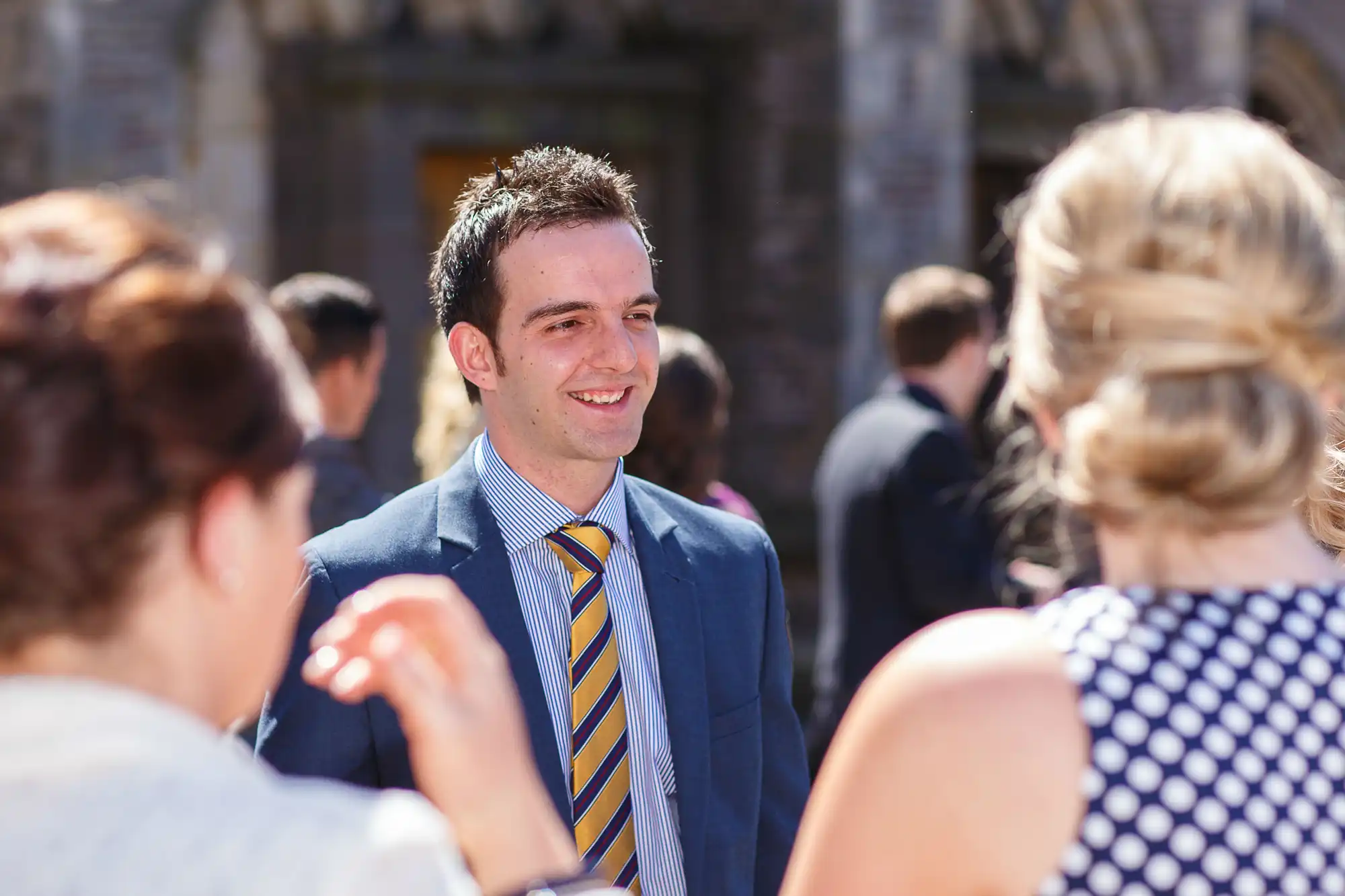 A man in a suit smiling while conversing with guests at a sunny, formal outdoor gathering.