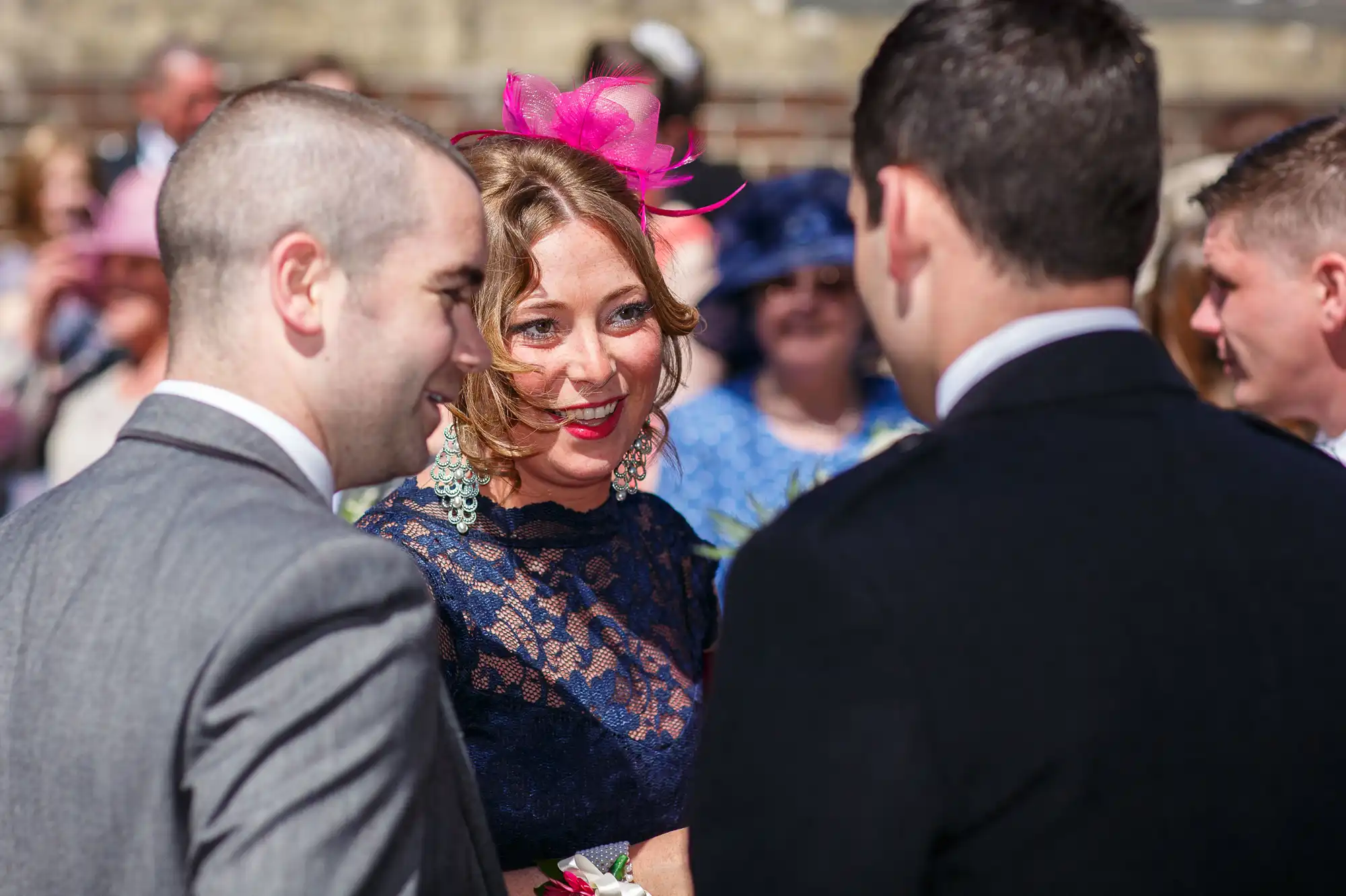 A woman in a blue lace dress and pink fascinator laughs while conversing with two men at a sunny outdoor social gathering.
