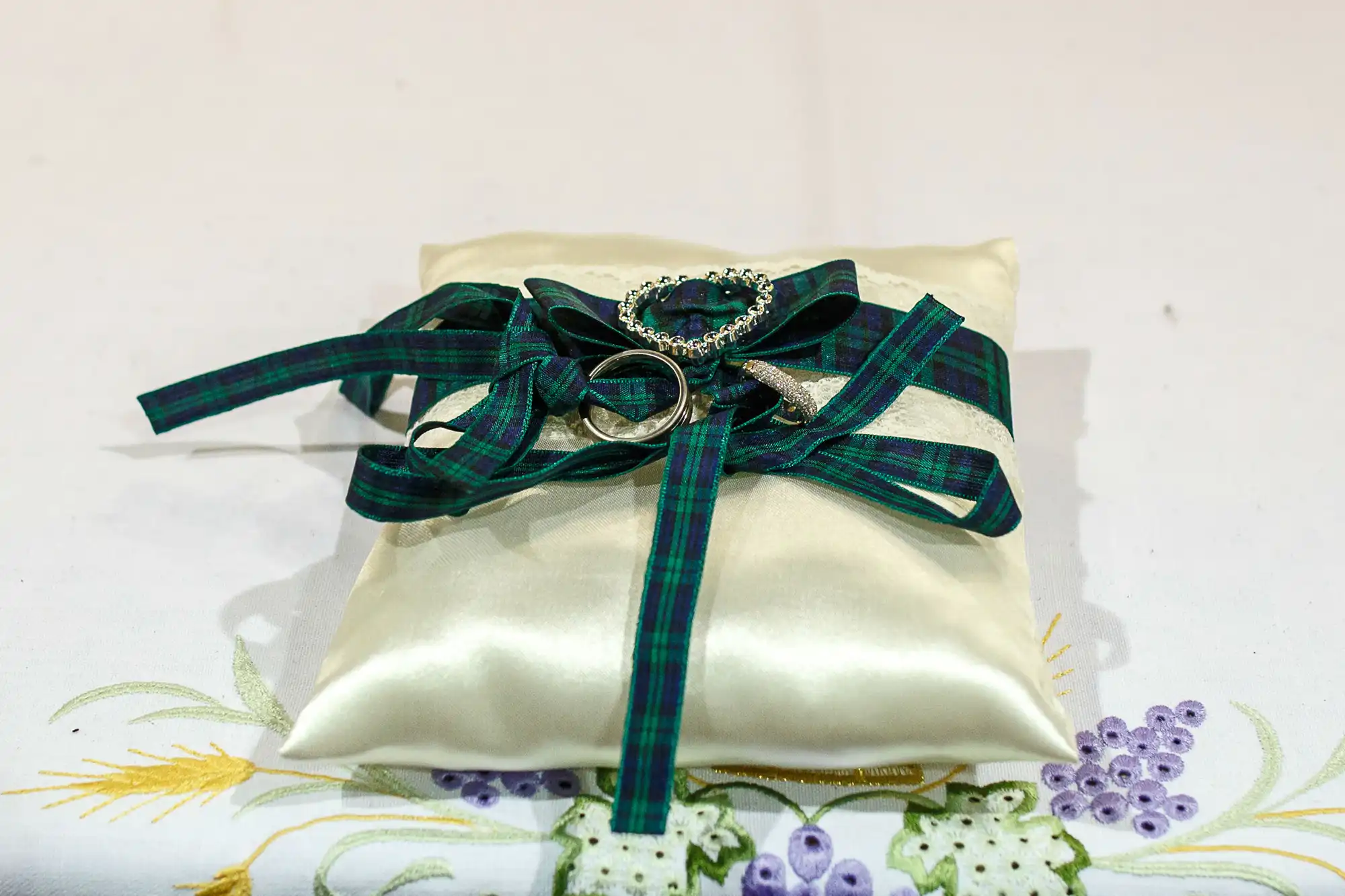 A satin ivory pillow adorned with a tartan ribbon and a sparkling brooch, typically used for holding rings at a wedding ceremony.