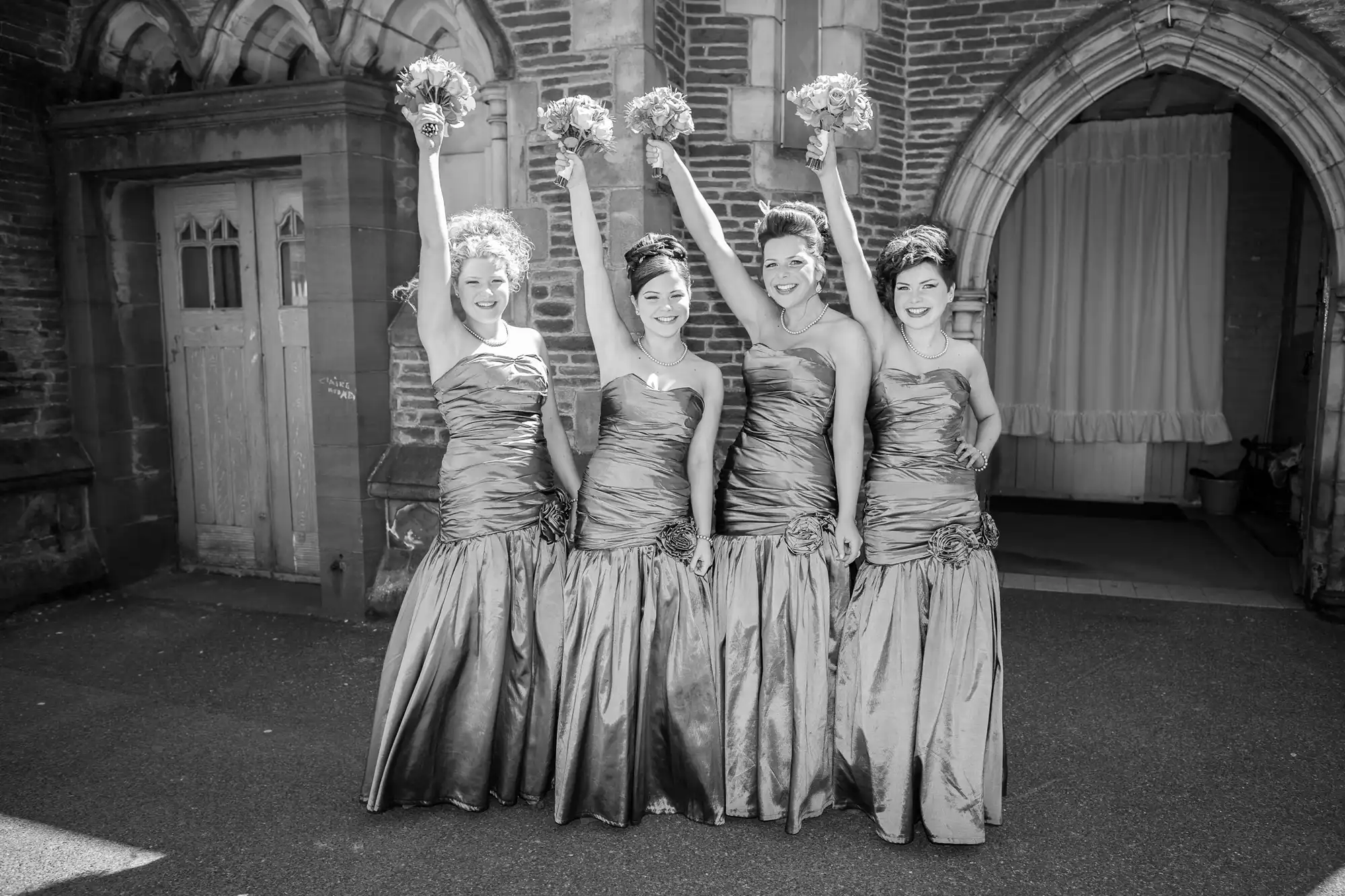 Four bridesmaids in matching dresses holding bouquets pose joyfully outside a church entrance.