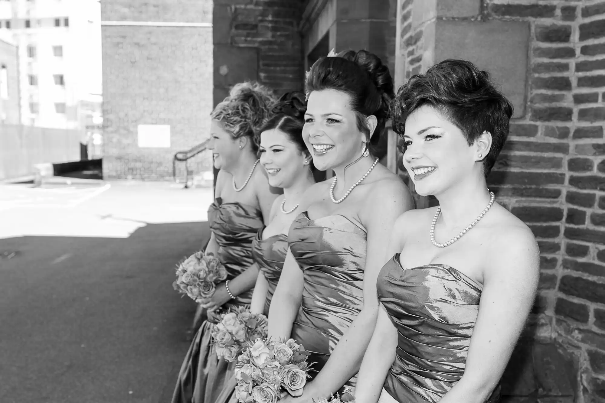 Four women in elegant dresses and pearl necklaces smiling outdoors at a wedding, black and white photo.