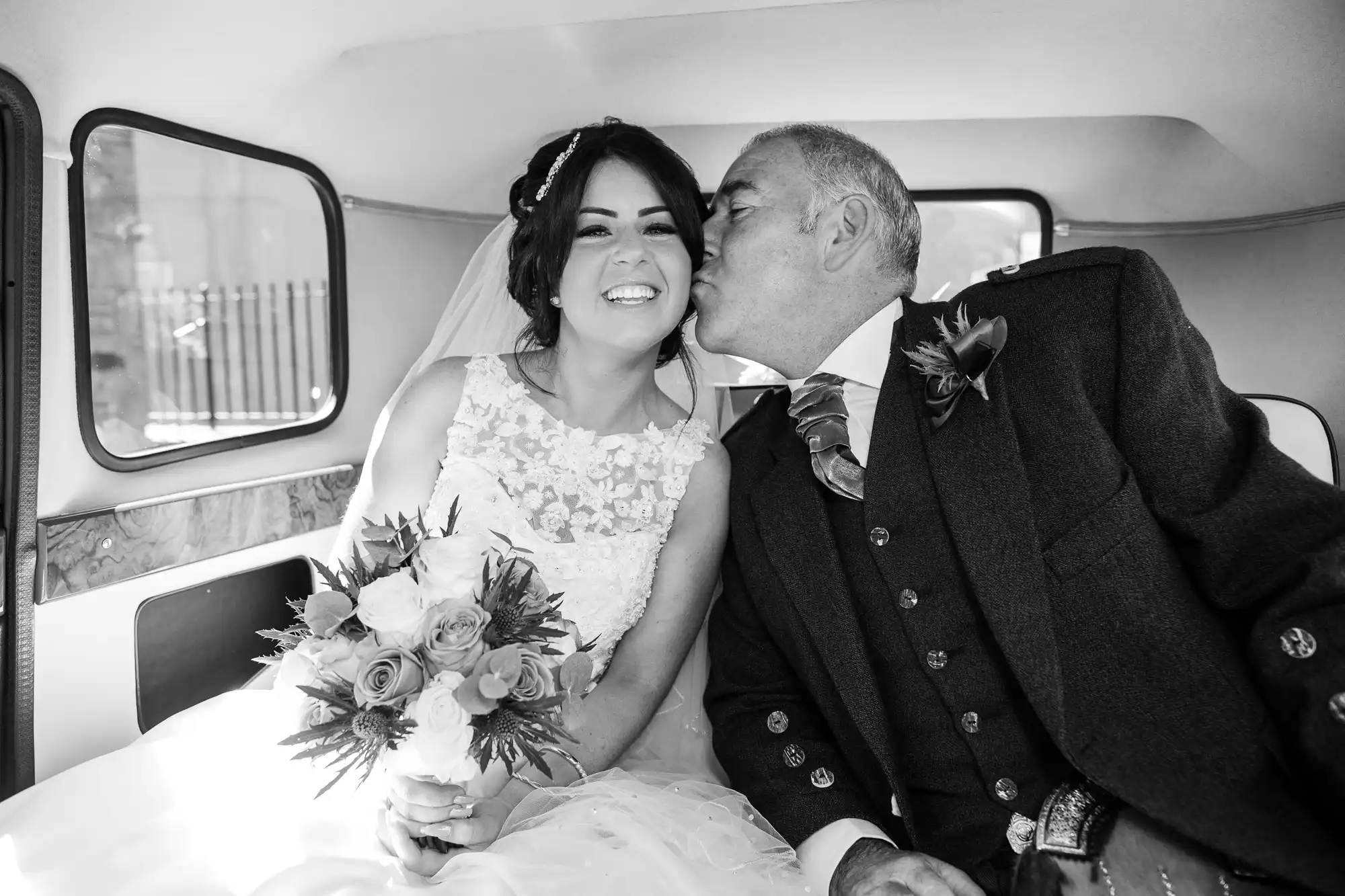 A bride in a white lace dress smiles joyfully as an older man in a traditional suit kisses her cheek inside a vintage car.