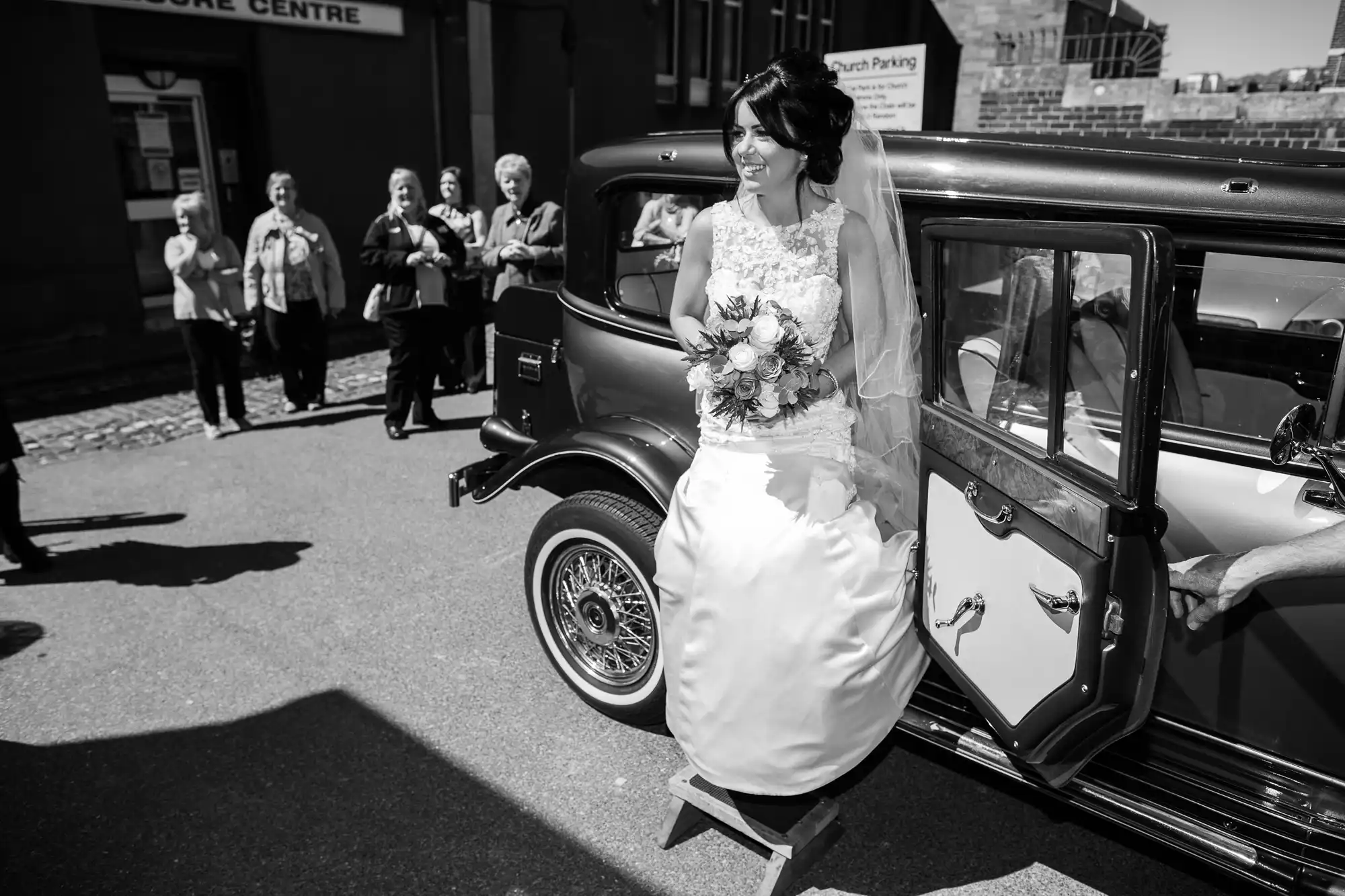 A bride stepping out of a vintage car, holding a bouquet, with onlookers in the background.
