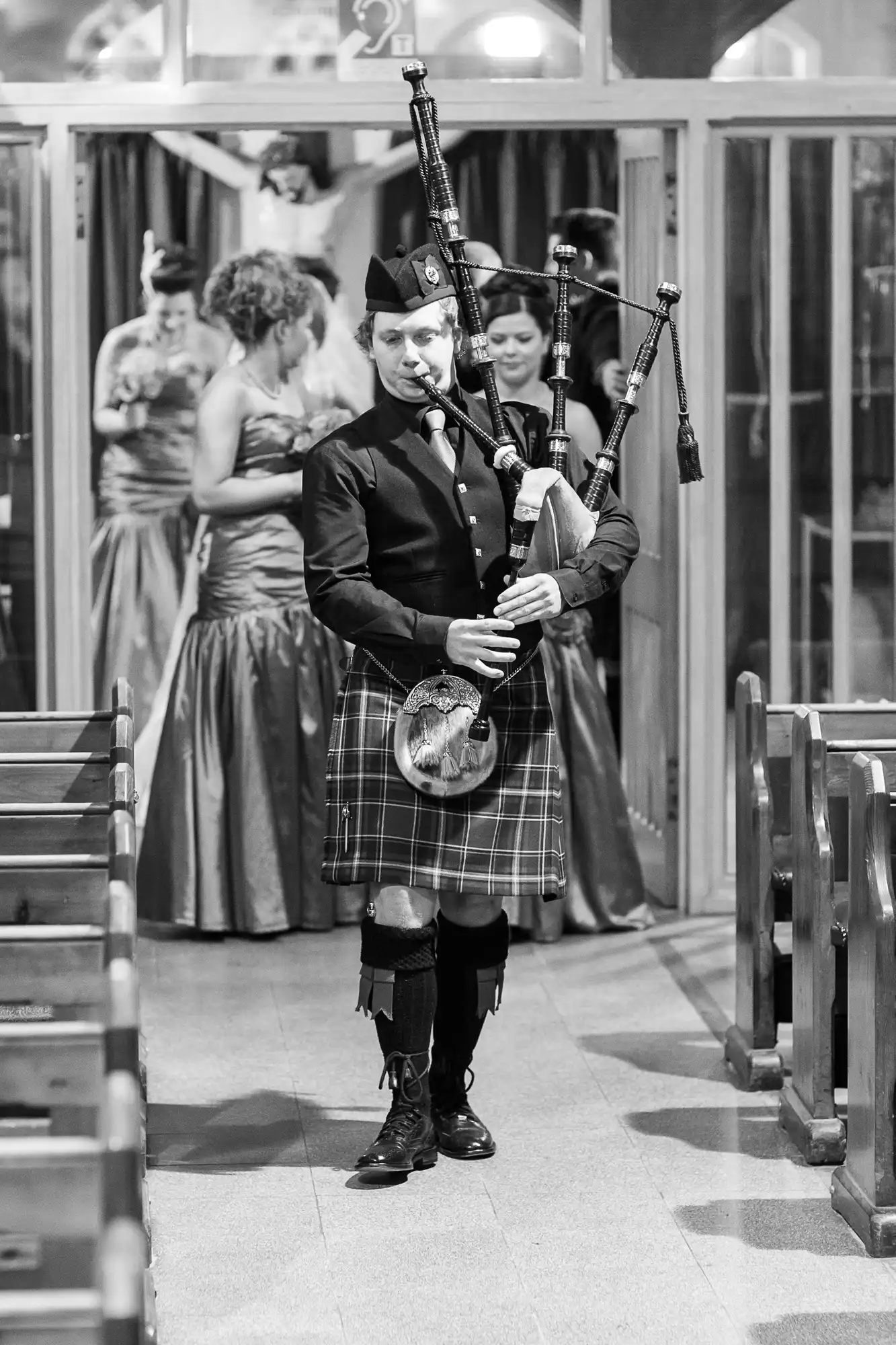 A bagpiper in traditional scottish attire plays inside a venue with people in the background.