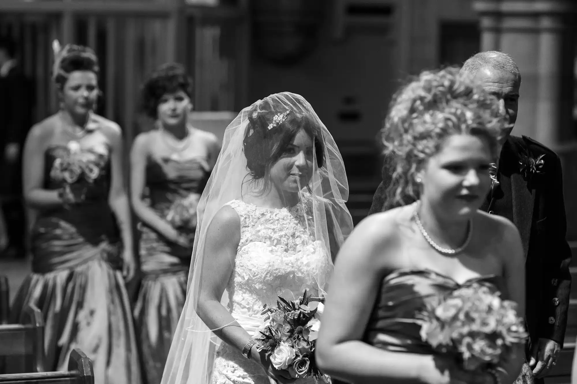 A black and white photo of a bride walking down the aisle, accompanied by guests in formal wear.