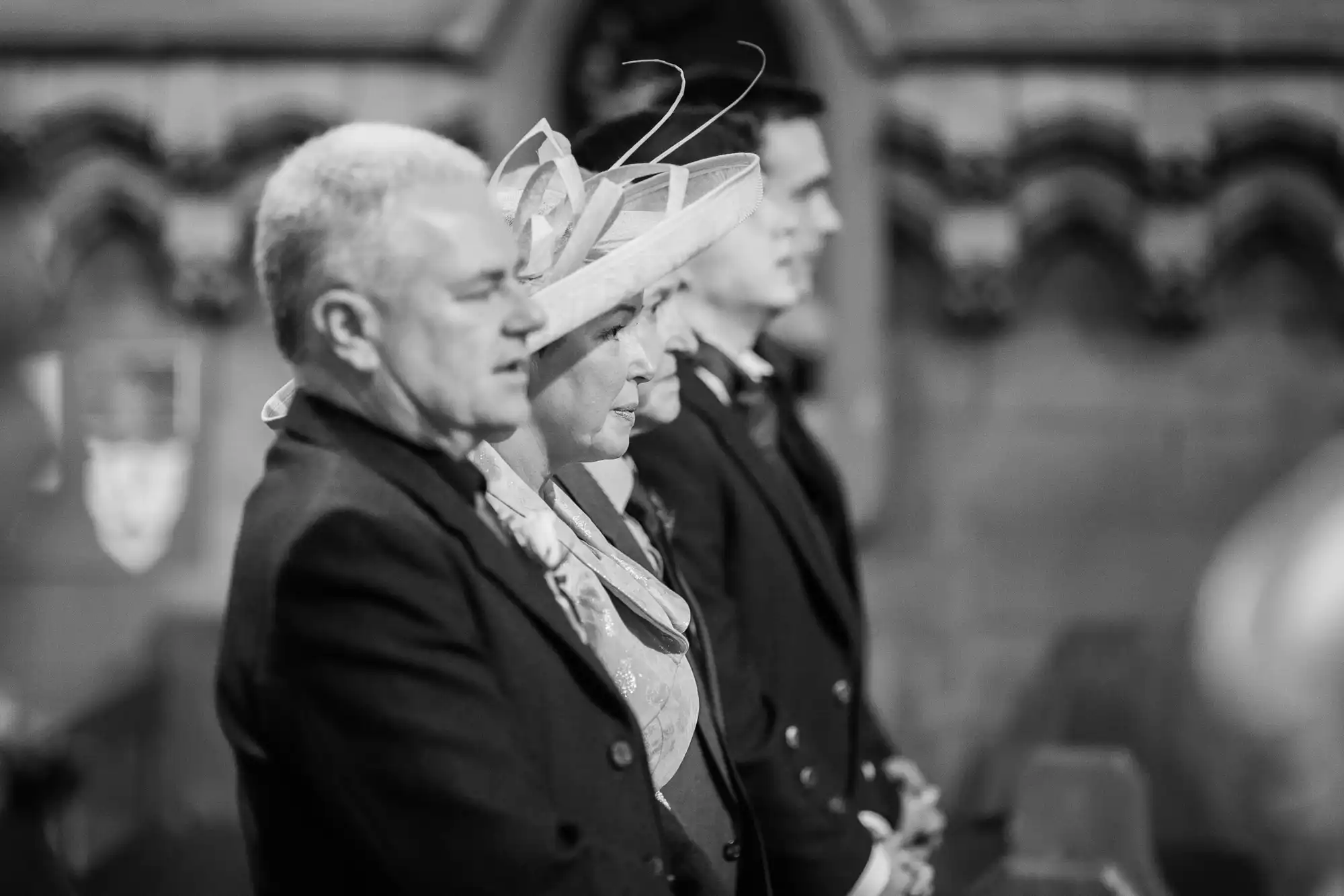 Three adults attending a formal event, standing solemnly side by side; the middle woman is wearing a large hat. the image is in black and white.