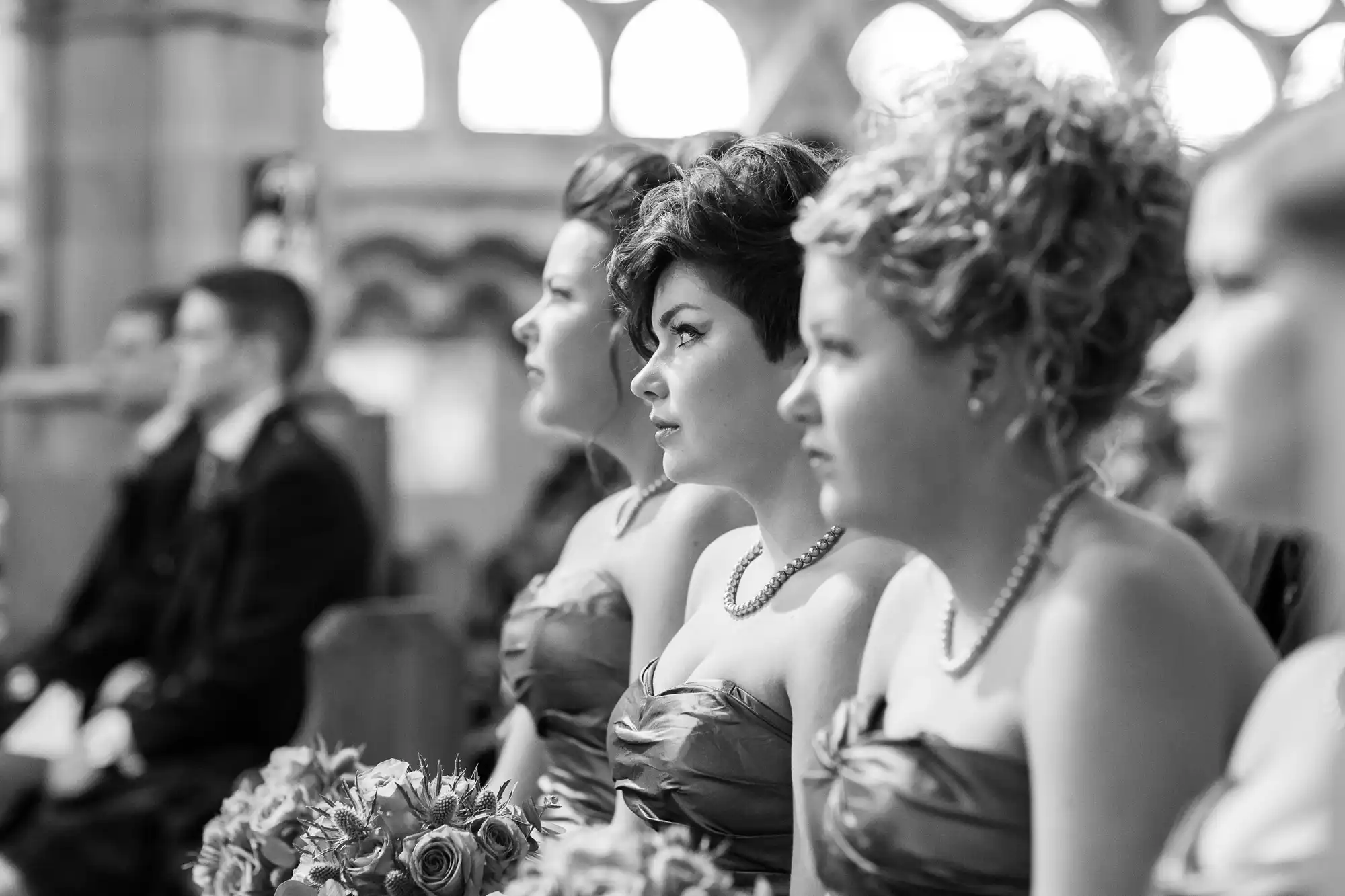 Black and white image of bridesmaids in elegant dresses, seated in a row at a wedding ceremony inside a church.