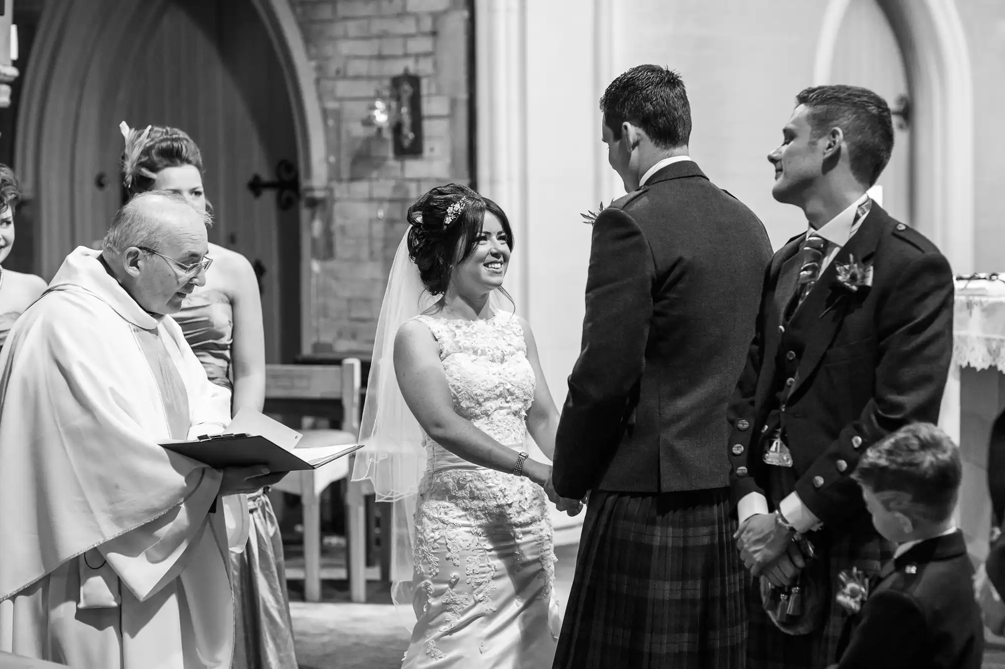 Bride in a lace dress smiling at the groom in a suit, with a priest and best man nearby, during a wedding ceremony inside a church.