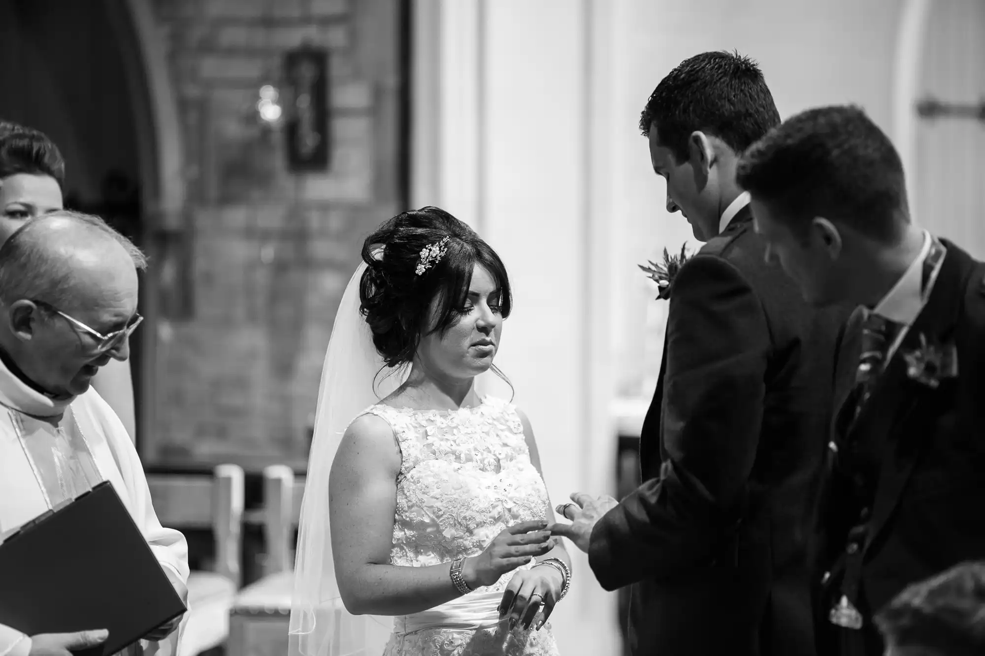 A bride and groom exchange rings during their wedding ceremony, officiated by a priest, inside a church. black and white image capturing a solemn yet joyful moment.