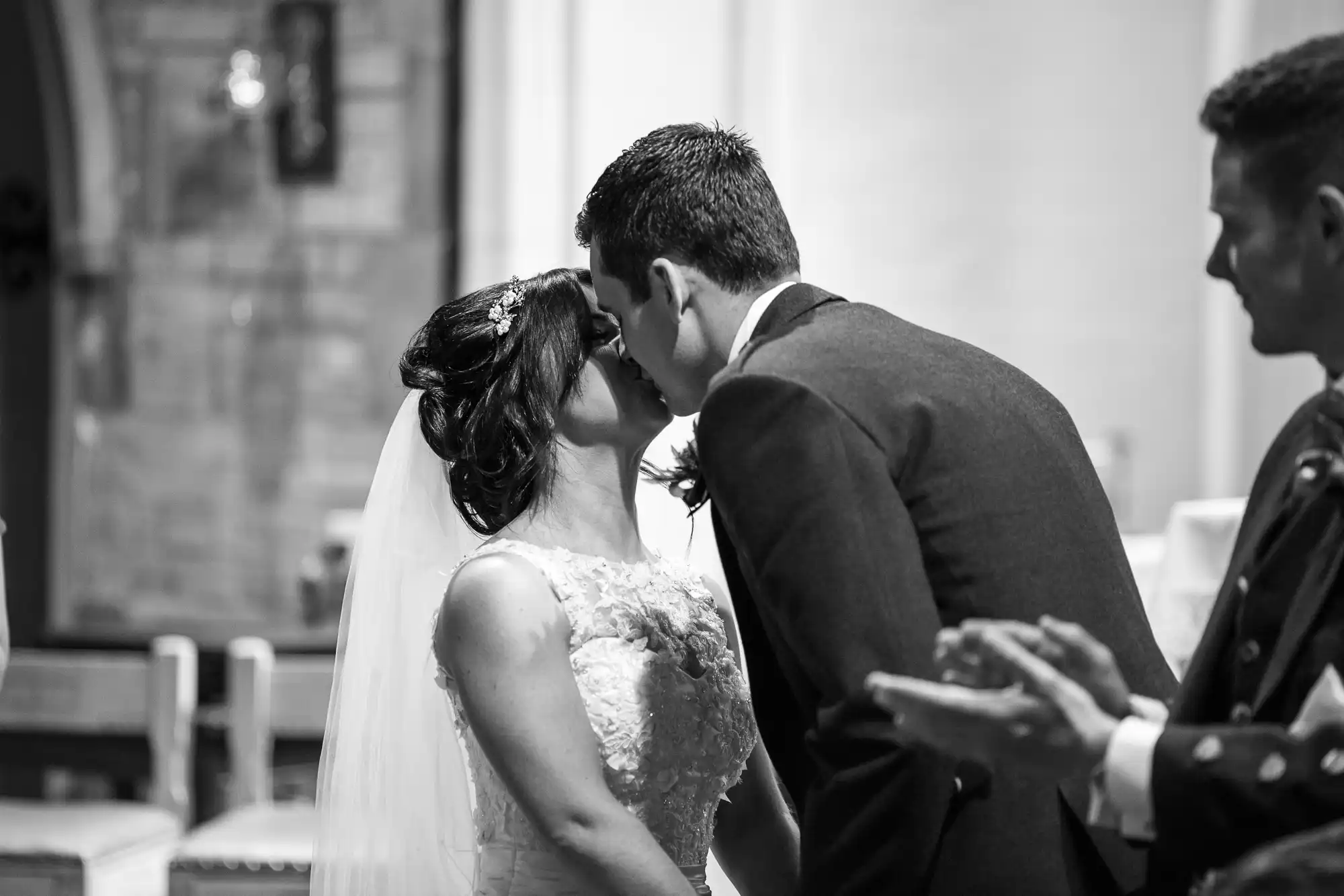 A bride and groom sharing a kiss at the altar during their wedding ceremony, in black and white, with an applauding guest visible.