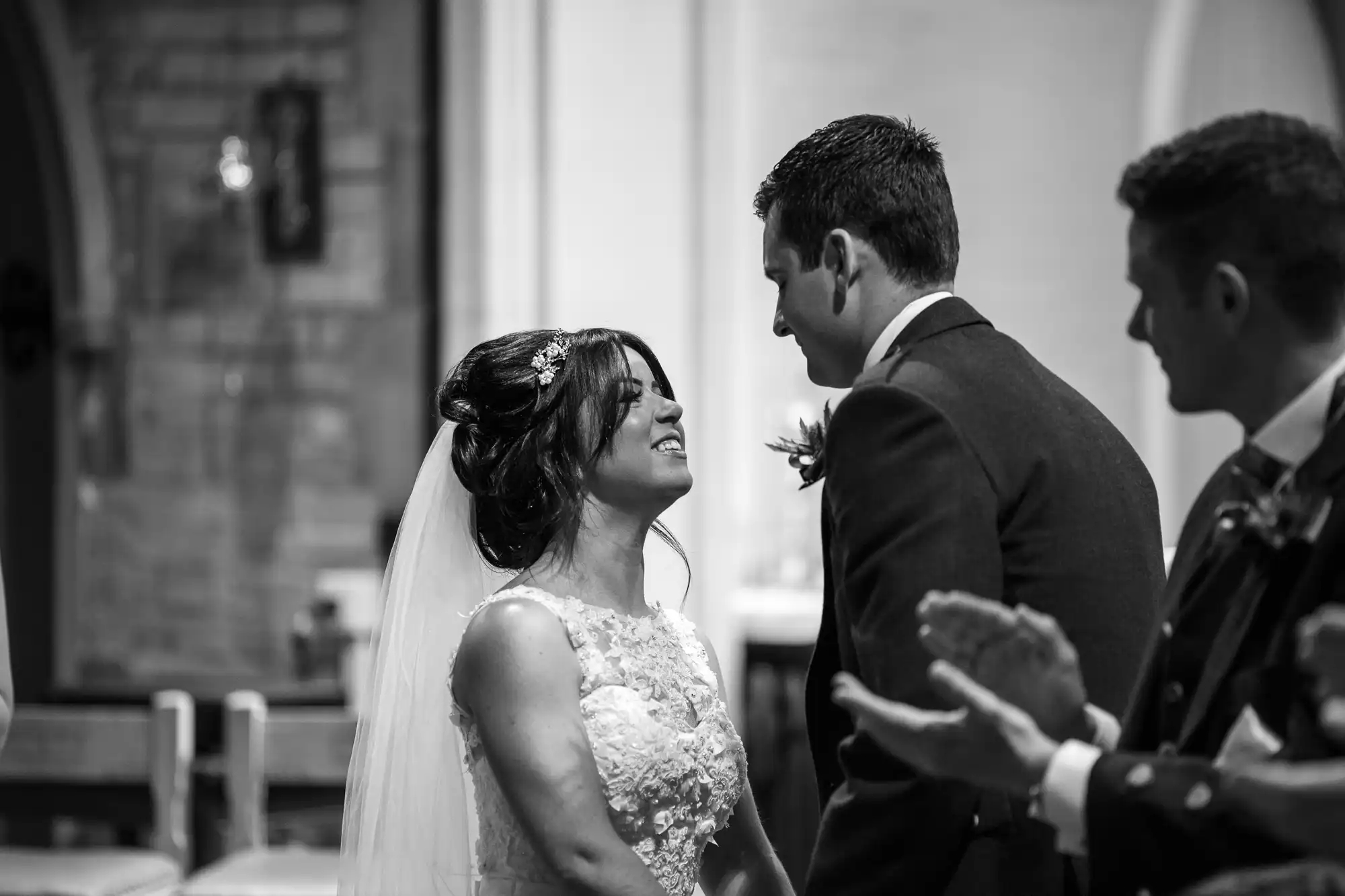 A bride and groom smiling at each other during their wedding ceremony in a church, captured in black and white.