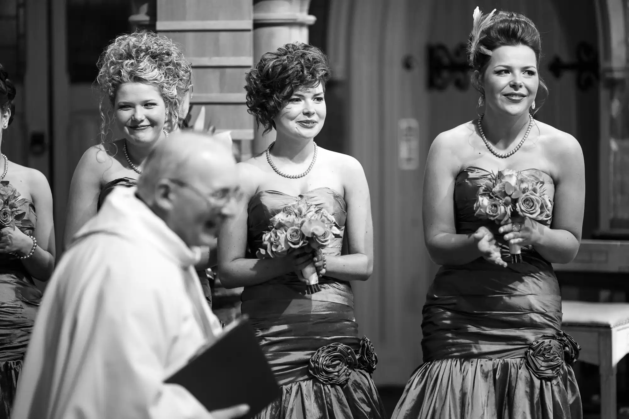 Three bridesmaids in elegant dresses with rosette details, smiling during a wedding ceremony in a black and white photo.