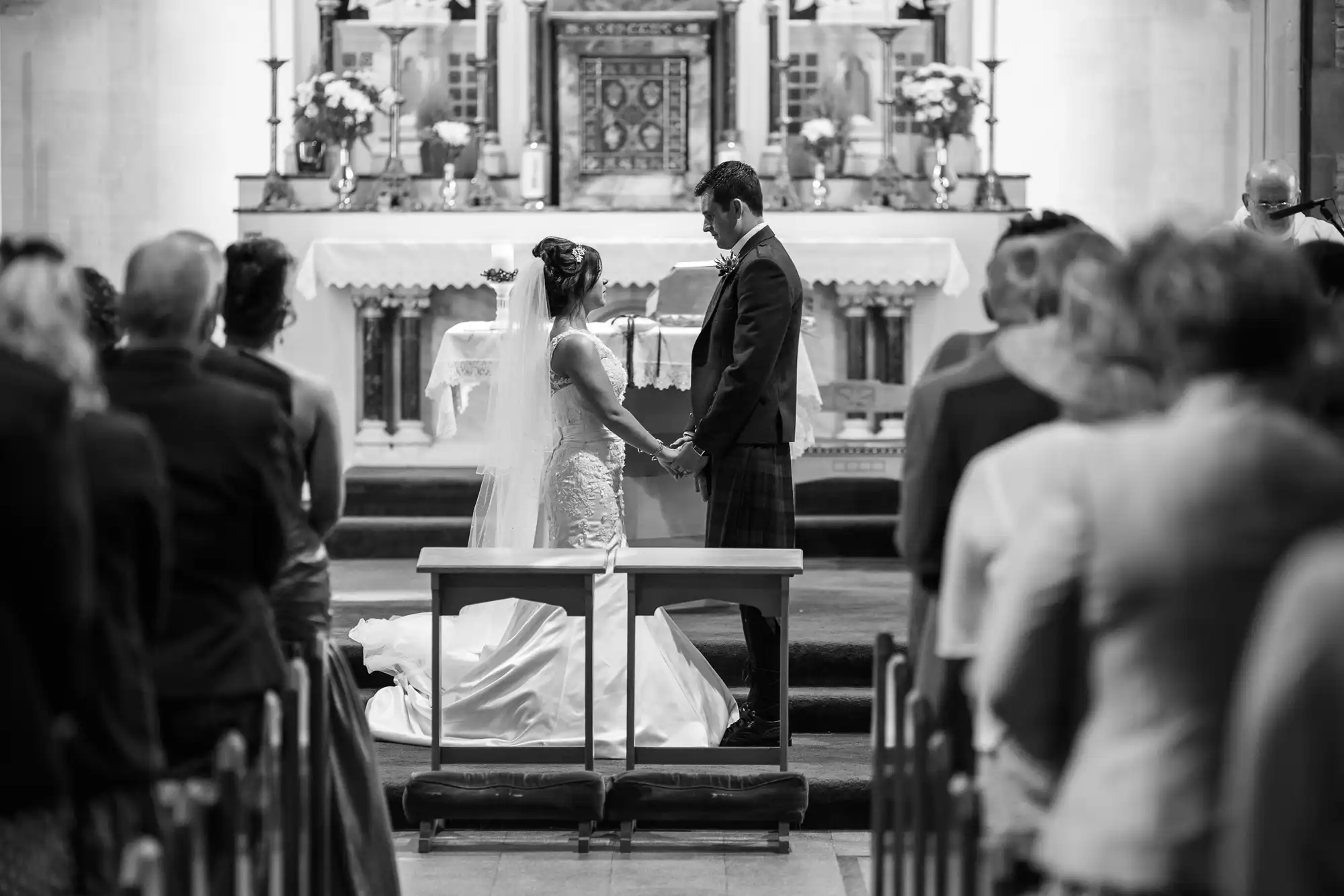 A bride and groom holding hands at the altar during a wedding ceremony in a church, viewed from behind the seated guests.