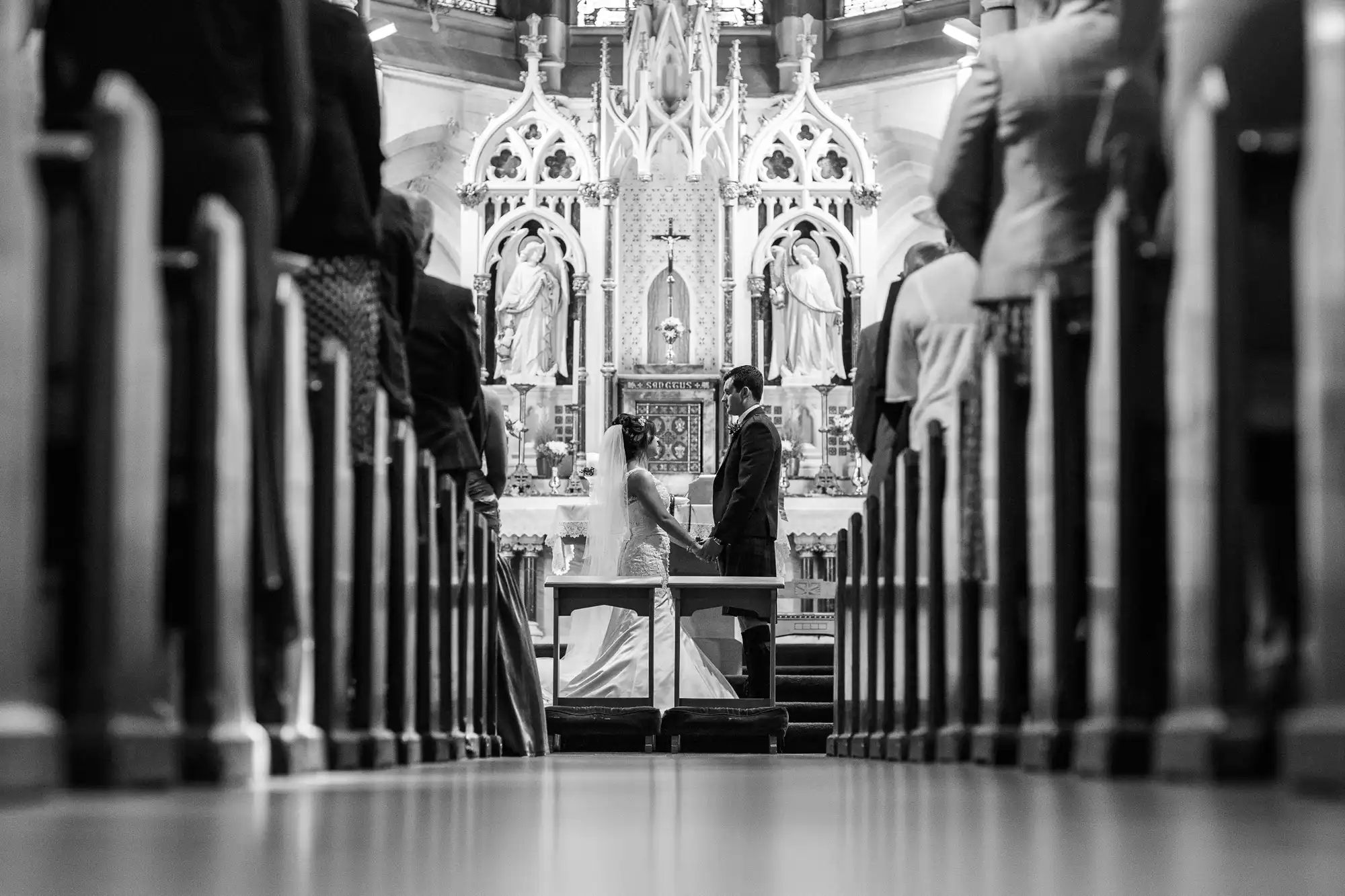 A bride and groom sit facing each other at an altar in a church, viewed between rows of guests' legs from the aisle.