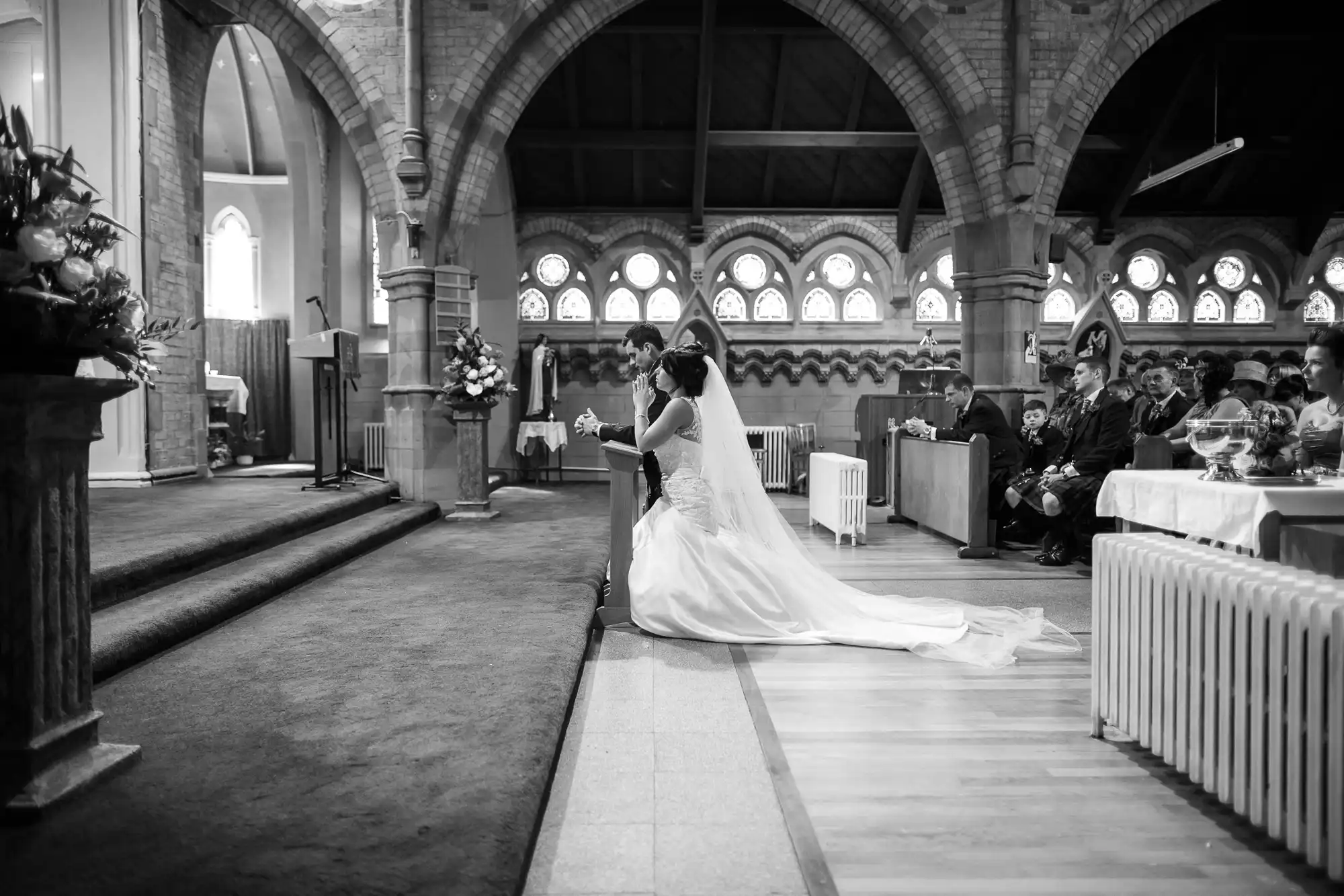A bride and groom kneel at the altar during a wedding ceremony in a church, with guests seated around them.