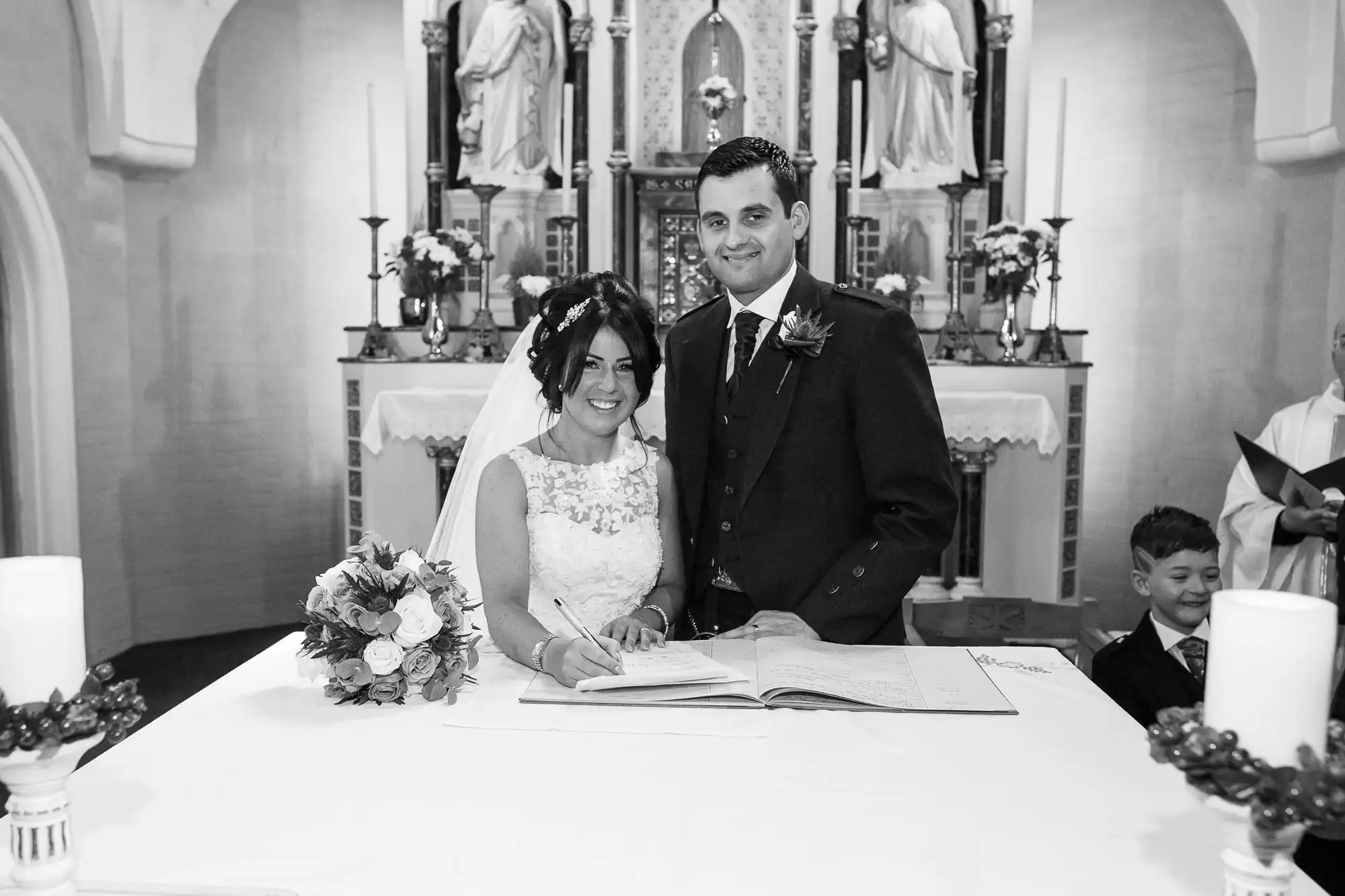 A bride and groom smiling and signing the marriage register in a church, with candles on the table and a priest behind them.