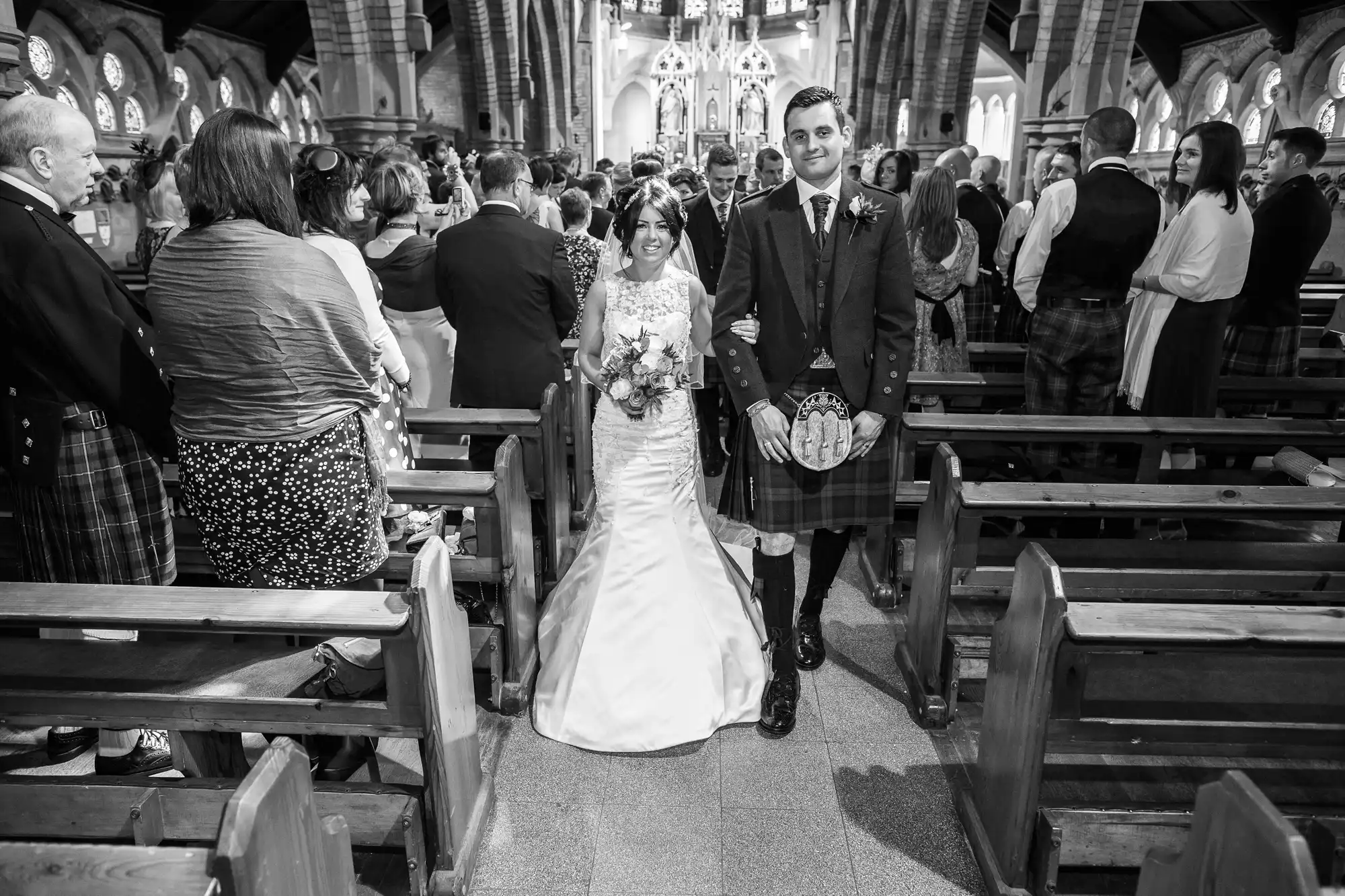 A bride and groom in a church, smiling as they walk down the aisle, surrounded by guests; the groom is wearing a kilt.