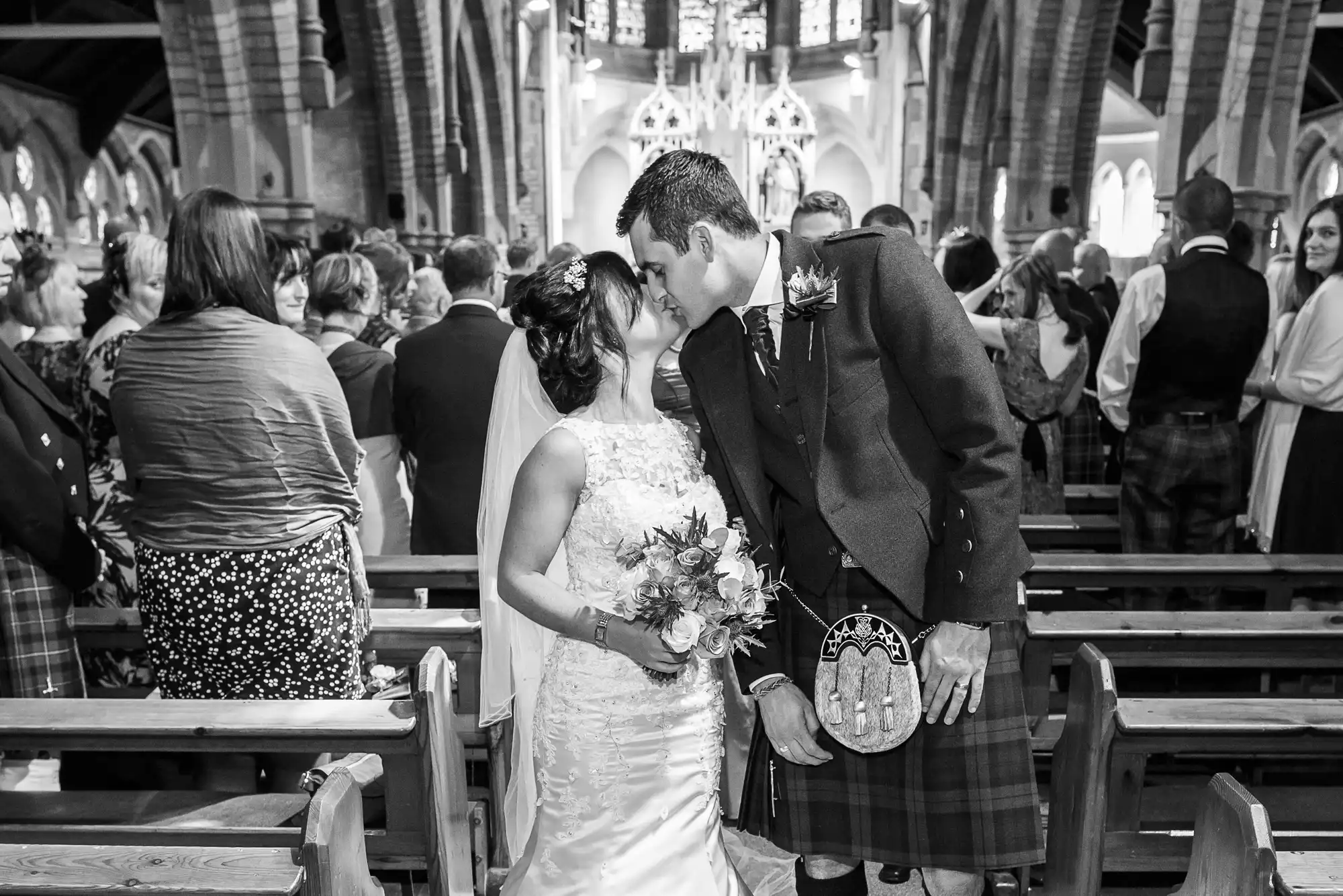 A bride and groom kissing in a church, surrounded by guests during their wedding ceremony, with the groom wearing a kilt.