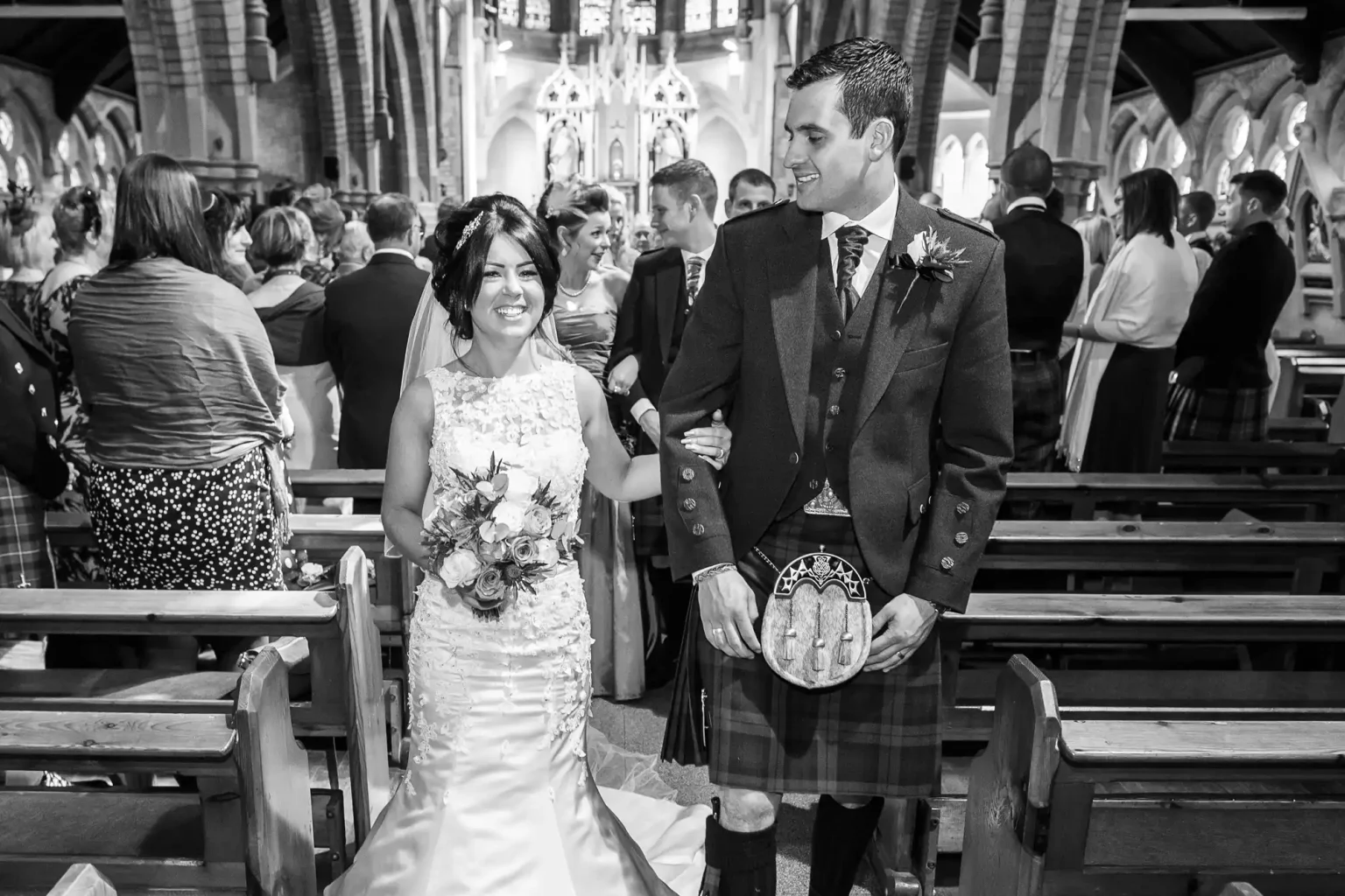 A bride in a white gown and a groom in a kilt smile as they walk down the aisle of a church filled with seated guests.