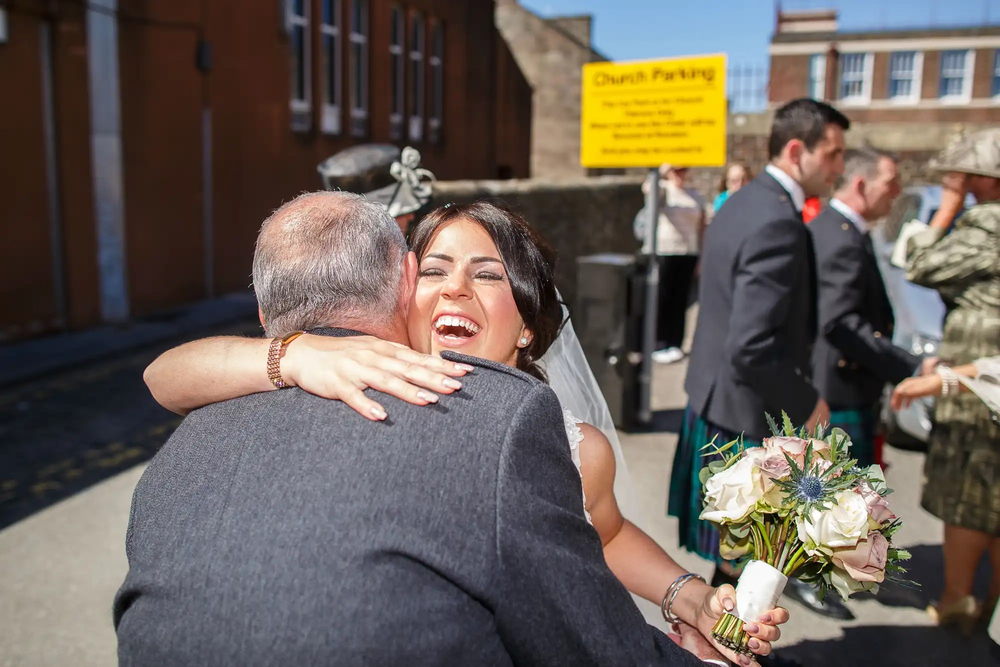 A bride joyfully hugs a guest outside a church on a sunny day, holding a bouquet, with other guests in the background.