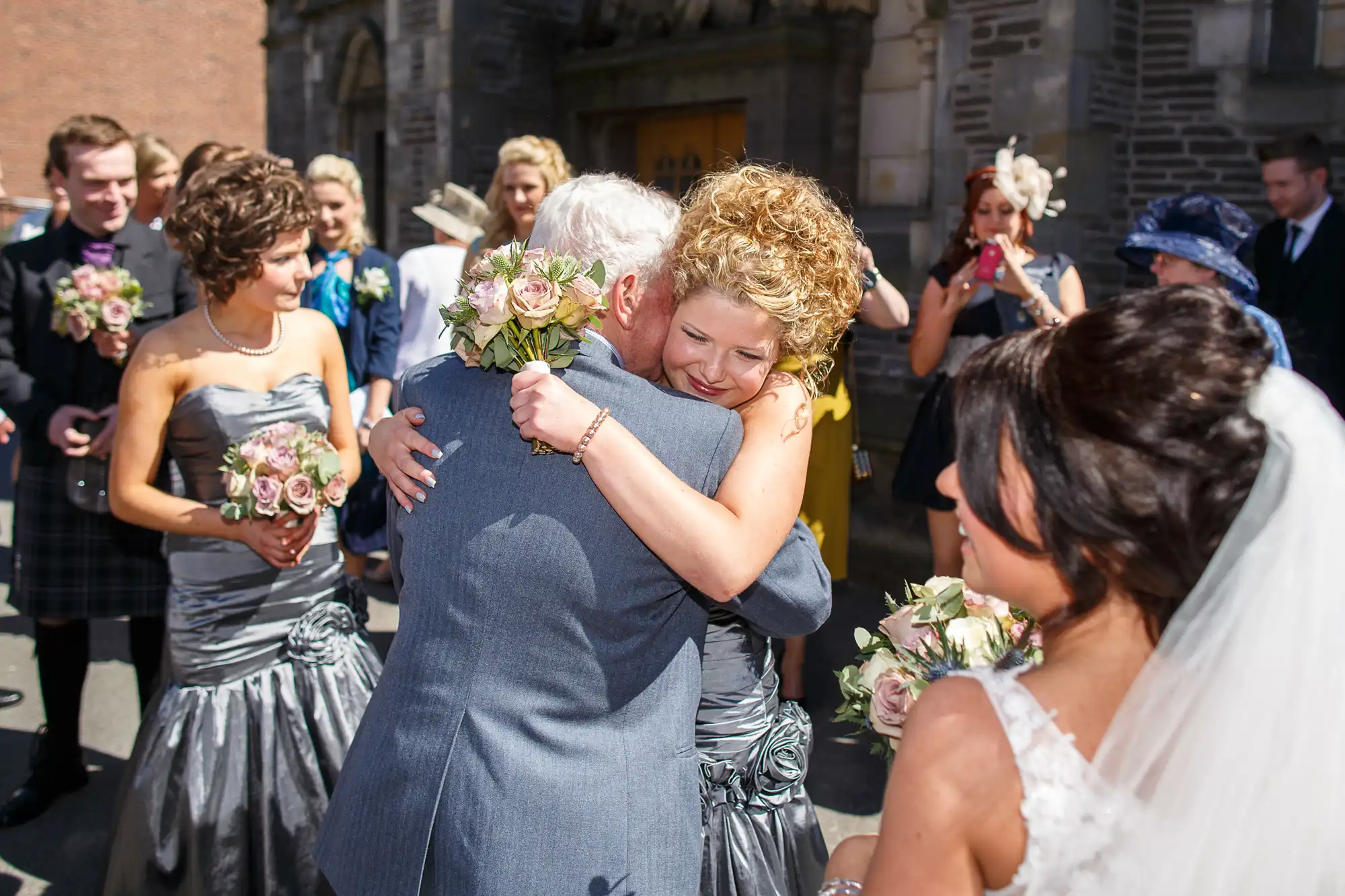 A joyful wedding scene where a woman hugs an older man amid guests, including a bride holding flowers on the right.