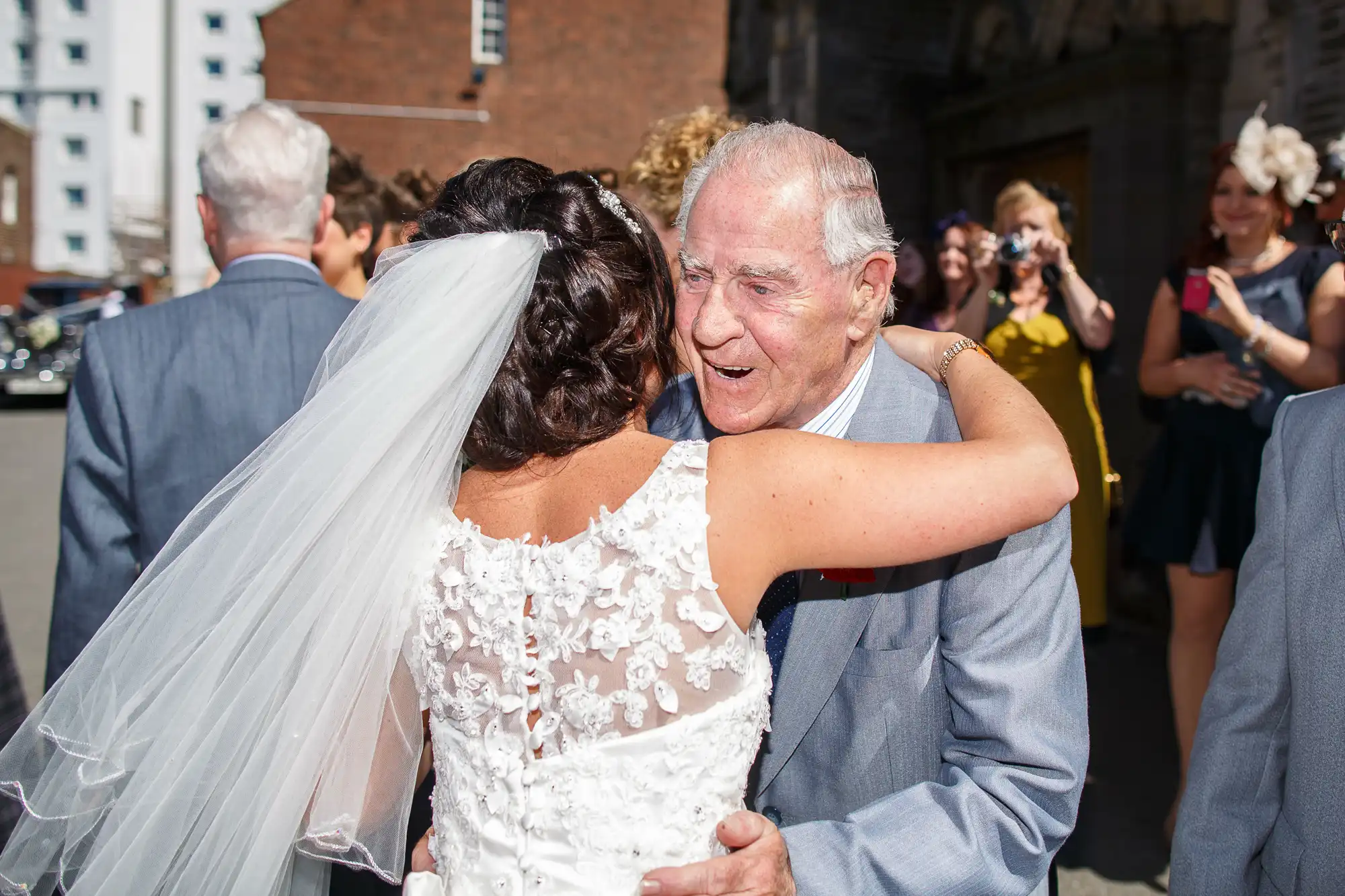 A bride in a white lace gown hugs an elderly man with joy at a sunny outdoor wedding ceremony, surrounded by guests.