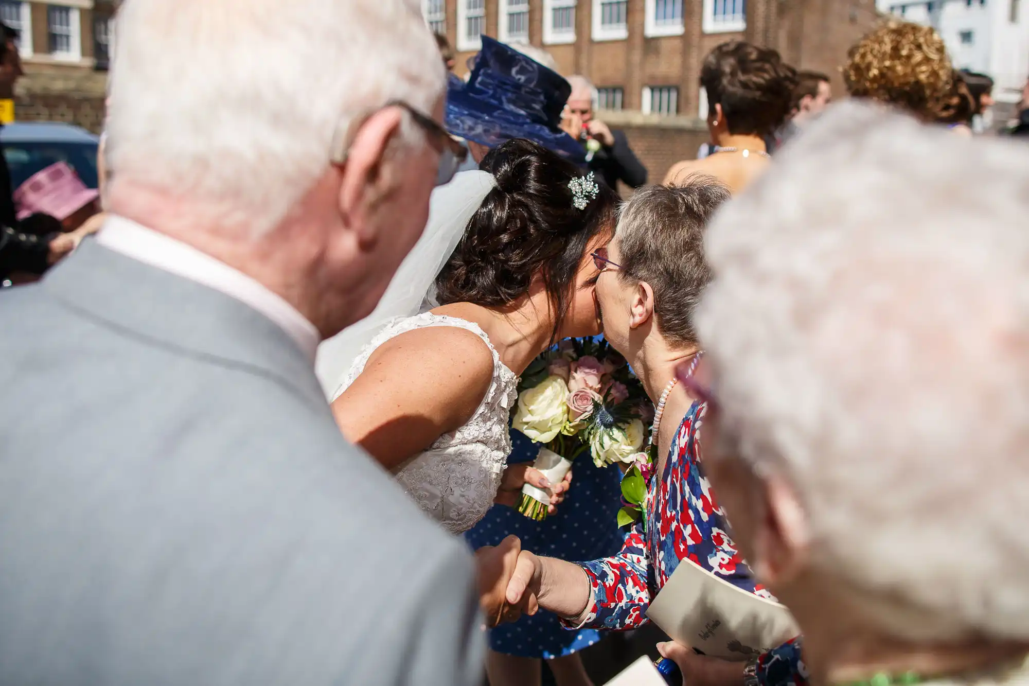 Bride in a white dress kissing an elderly woman's cheek outside; guests in formal attire surround them.