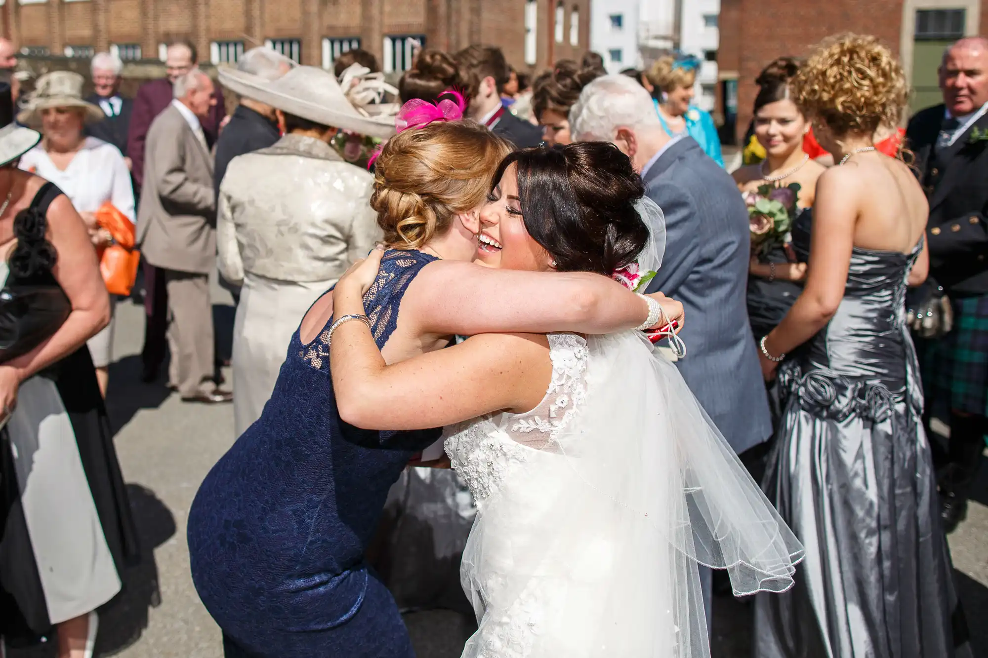 A bride in a white dress hugs a woman in a blue dress at a sunny outdoor wedding reception, surrounded by guests in formal attire.