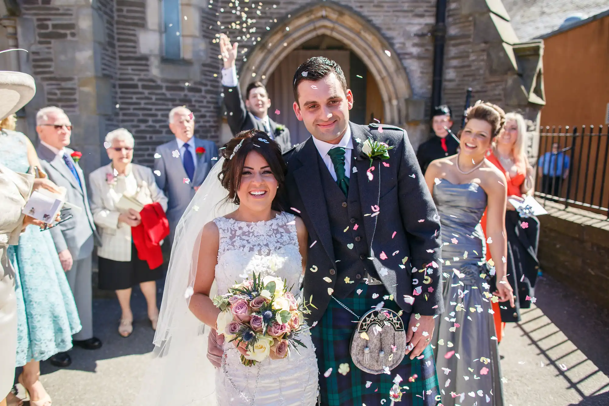 A bride and groom smiling outside a church as guests throw confetti over them on a sunny day.