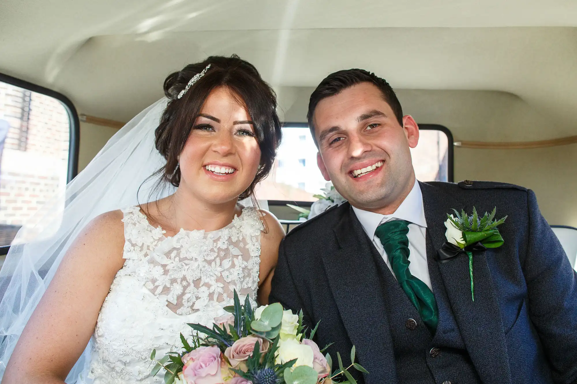 A newlywed couple smiling inside a car, the bride in a lace dress holding a bouquet, and the groom in a green tie and suit with a floral boutonniere.