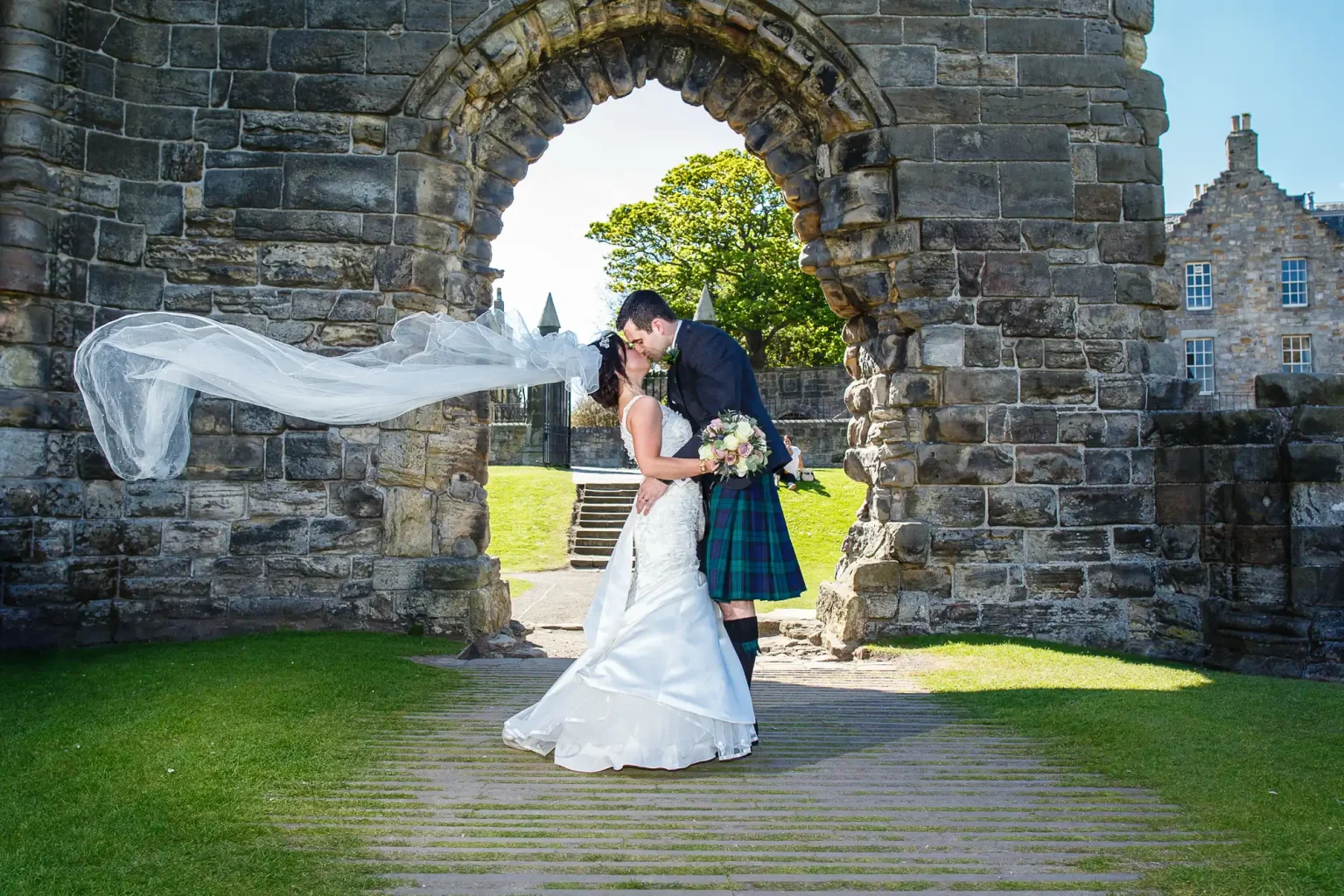 Fairmont St Andrews wedding photographer image of a bride and groom kiss under a stone archway; the bride's veil is blowing in the wind, and the groom is wearing a kilt.