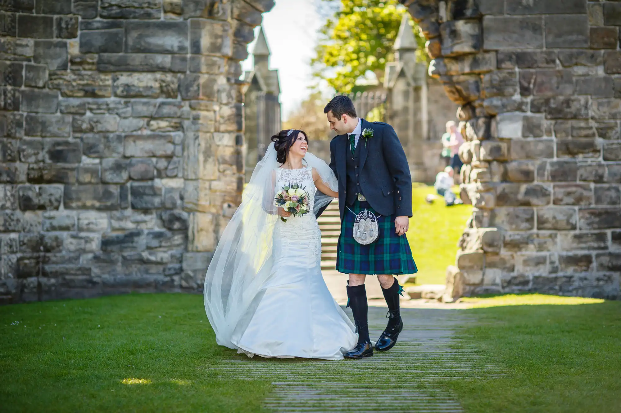 Bride in a white dress and groom in a tartan kilt smiling at each other on a grassy path, with historical stone arches in the background.