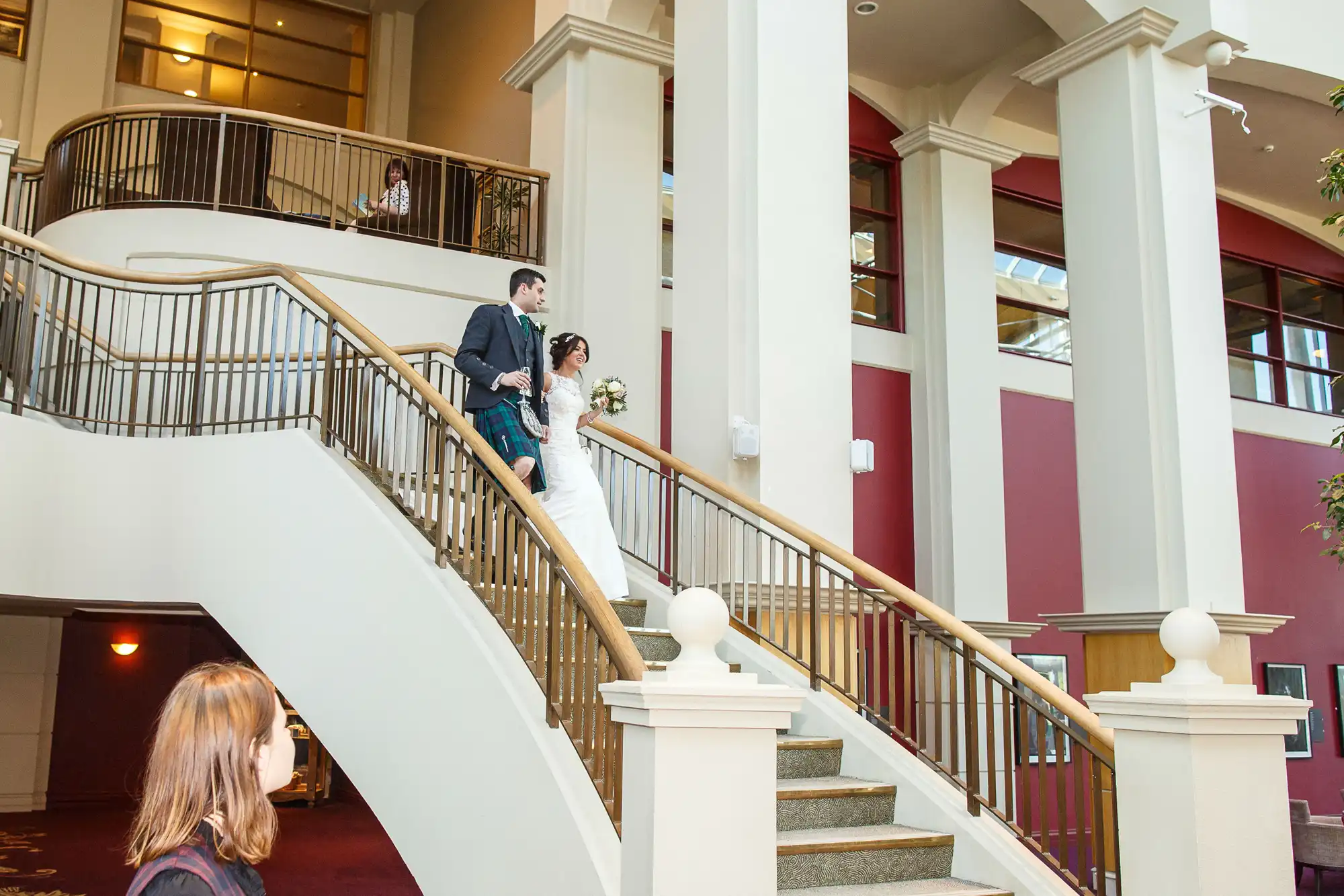 A bride and groom descending a curved staircase in a grand hall, with a guest watching from below.