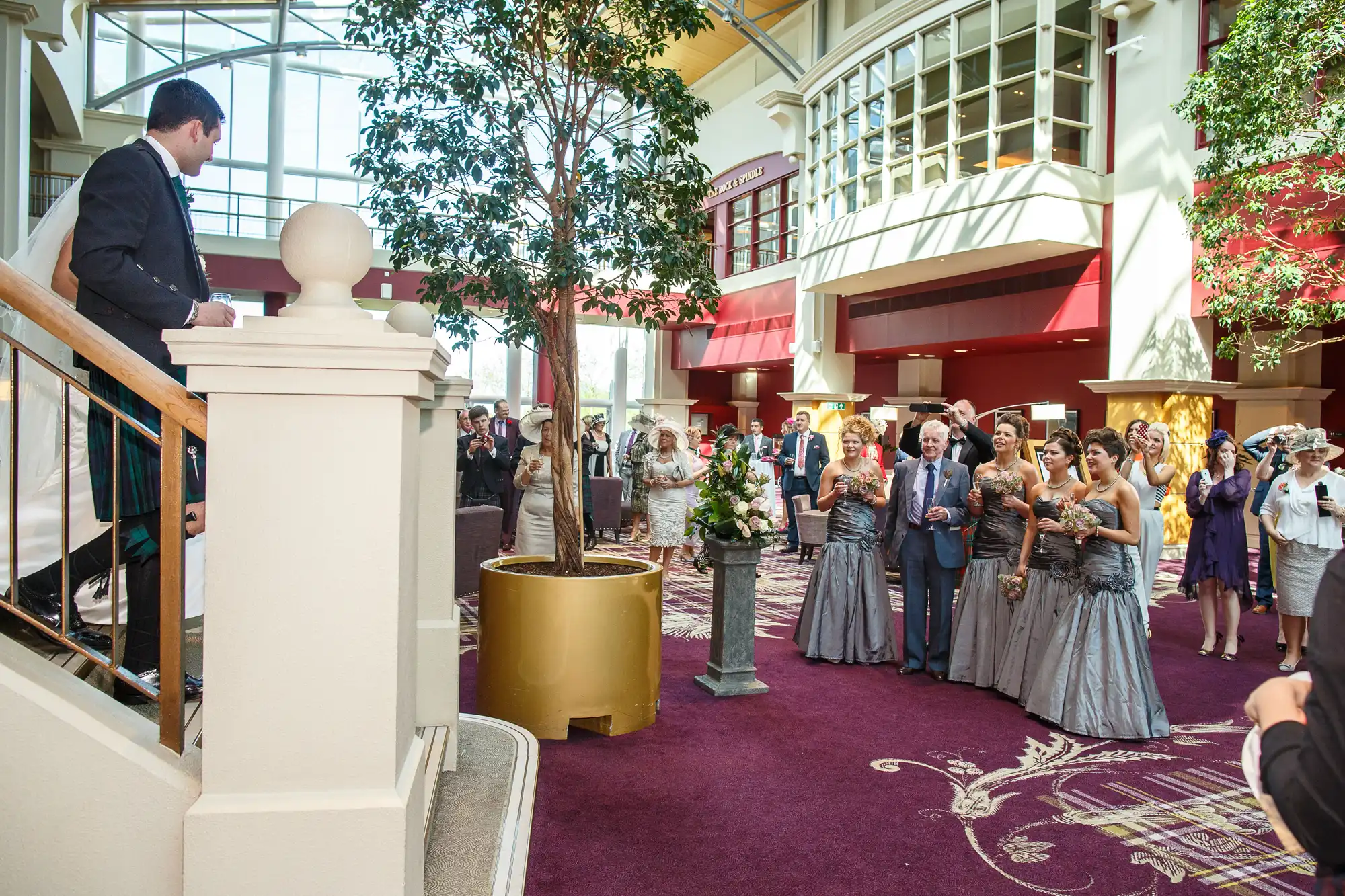 A wedding ceremony in an indoor atrium with guests watching a bride walking up stairs, and a groom waiting by a railing.