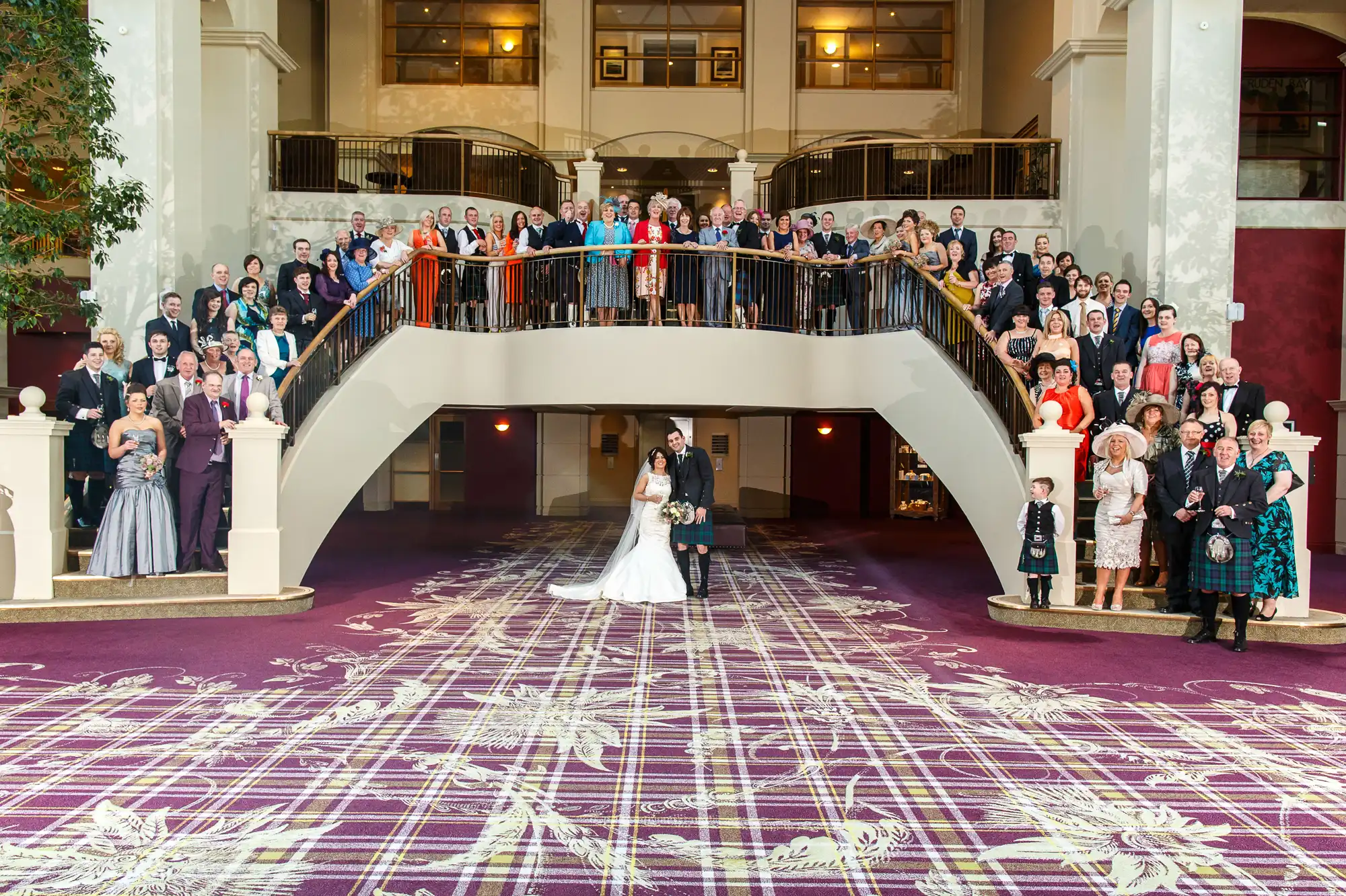 A large wedding group photo in a grand hotel lobby with the bride and groom centered on the floor, guests assembled on surrounding staircases and balcony.