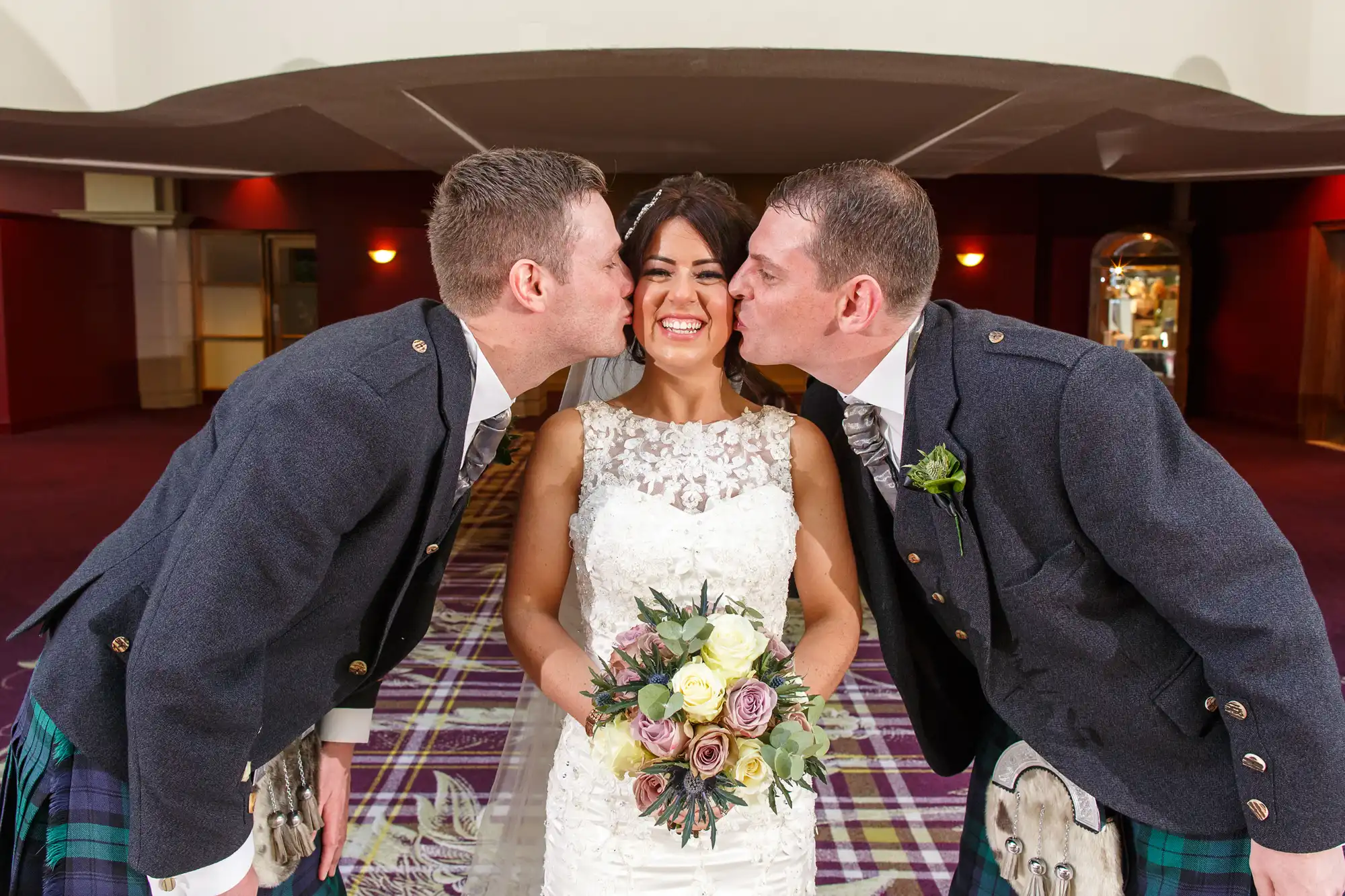 A bride holding a bouquet laughs as she is kissed on each cheek by two men in kilts and blazers at a wedding venue.