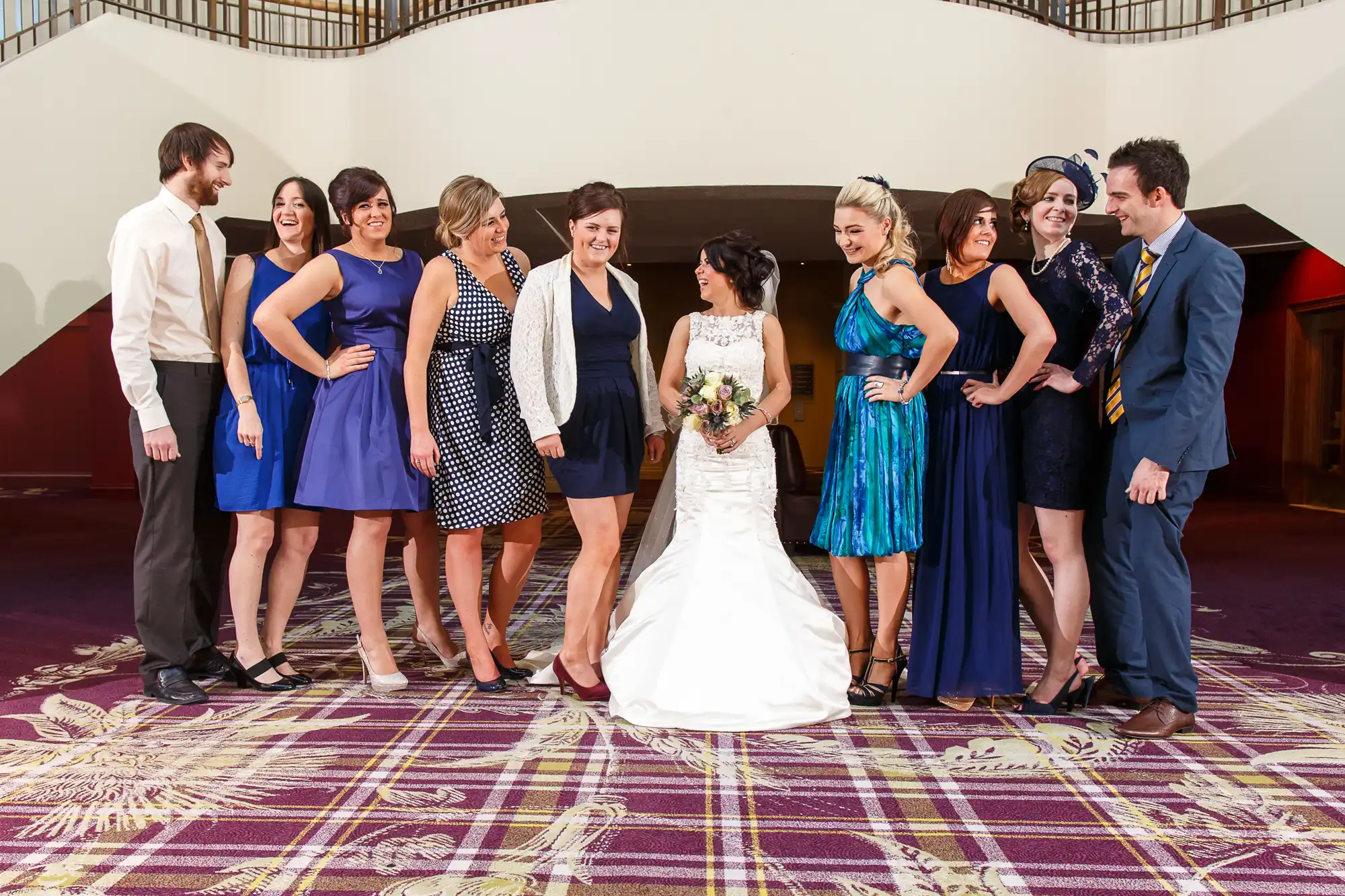 A bride in a white gown and groom in a navy suit with a group of guests in semi-formal attire, smiling and posing in an indoor setting.